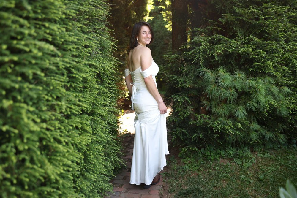 Bride in white off-shoulder wedding gown walking along a lush green garden pathway.