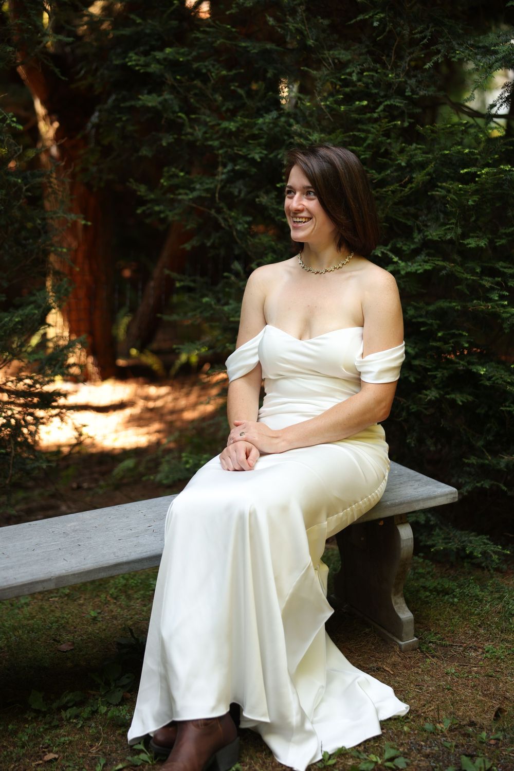 Bride in white off-shoulder gown sitting on a stone bench surrounded by green trees.