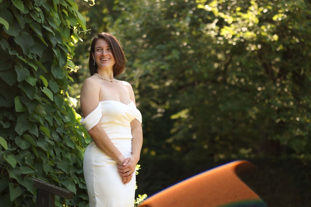 Bride in white off-shoulder wedding dress smiling beside ivy-covered wall in garden.