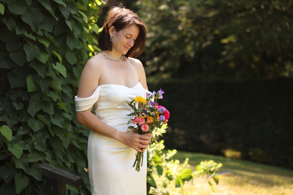 Bride in white off-shoulder gown holding colorful bridal bouquet in sunlit garden.