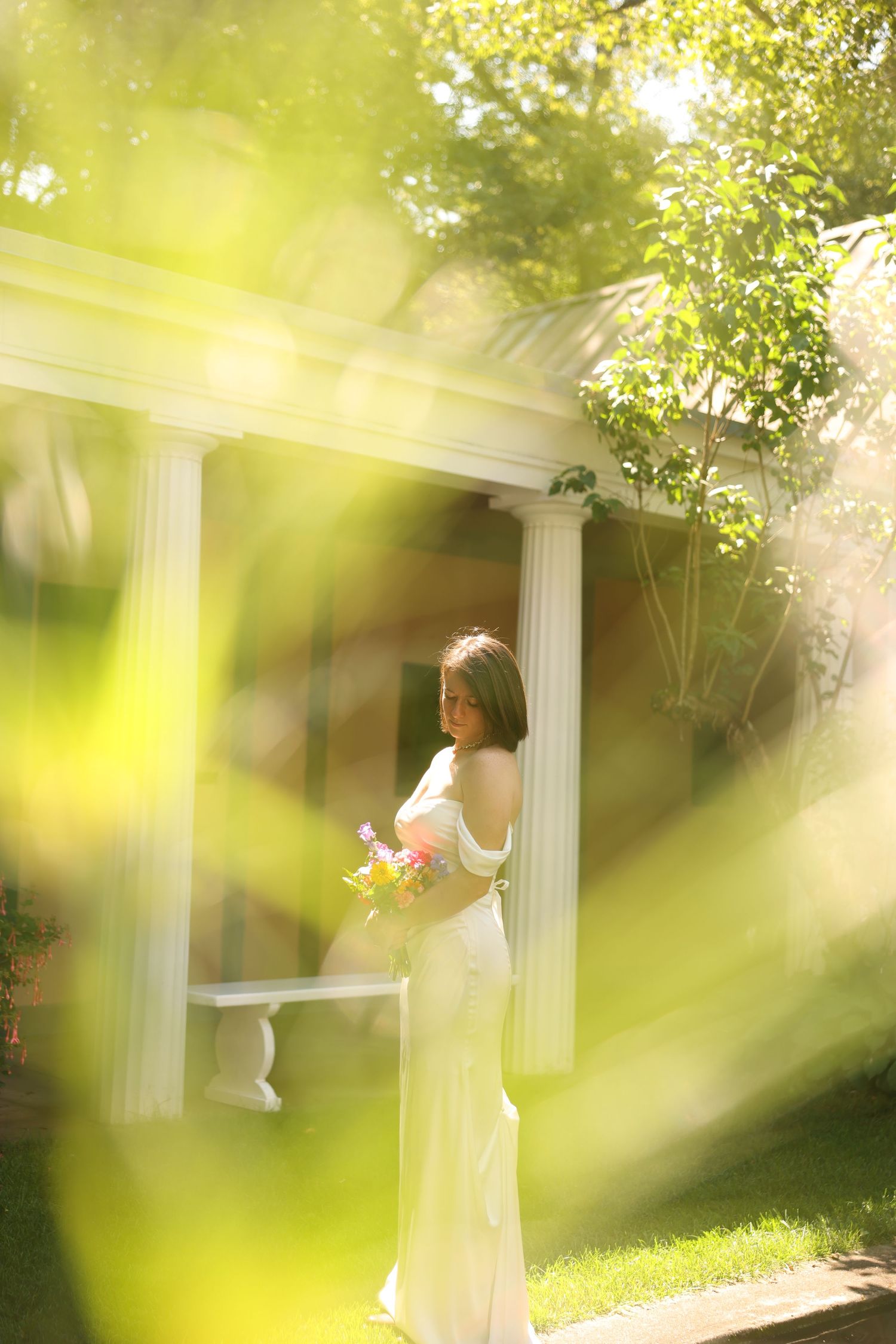 Bride in white gown holding colorful bouquet standing by white pillared gazebo in sunlight.