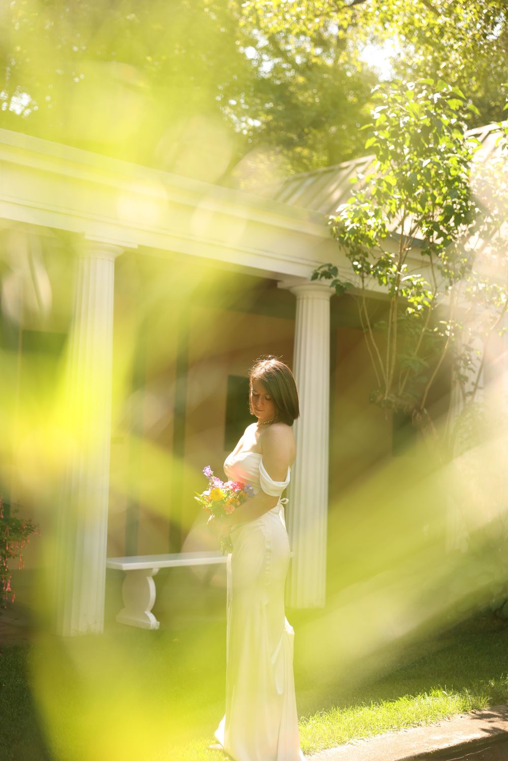 Bride in white gown holding colorful bouquet standing by white pillared gazebo in sunlight.