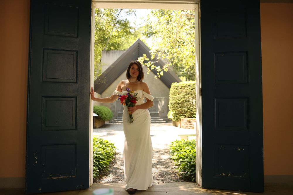 Bride in white off-shoulder gown holding bouquet framed by large open dark wooden doors.