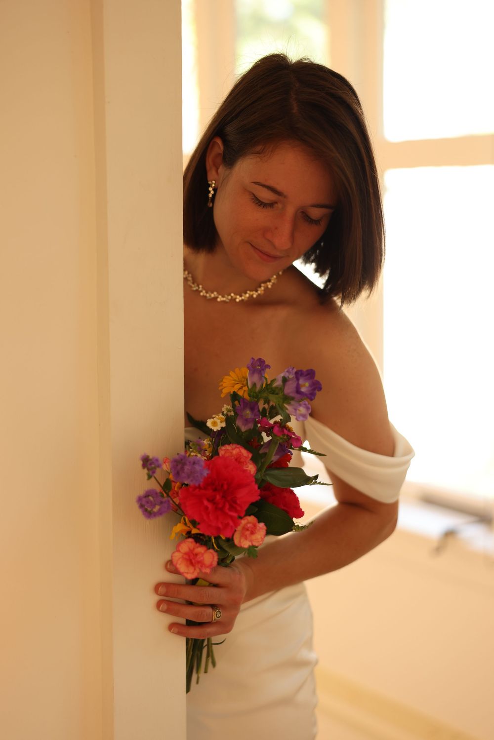 Bride in white off-shoulder gown holding colorful floral bouquet beside an interior column.