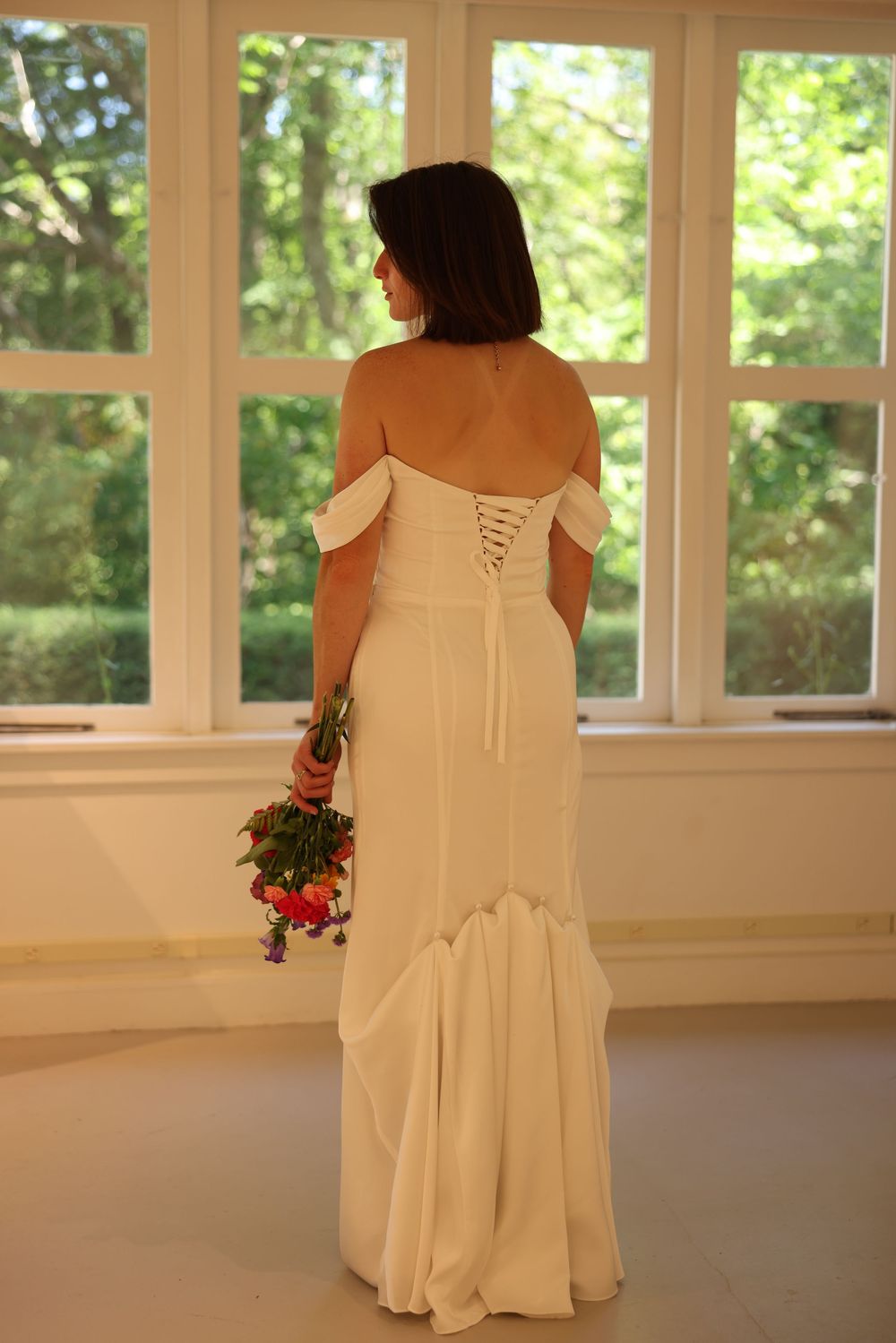 Bride in white off-shoulder corset-back gown holding bouquet gazing out large windows.