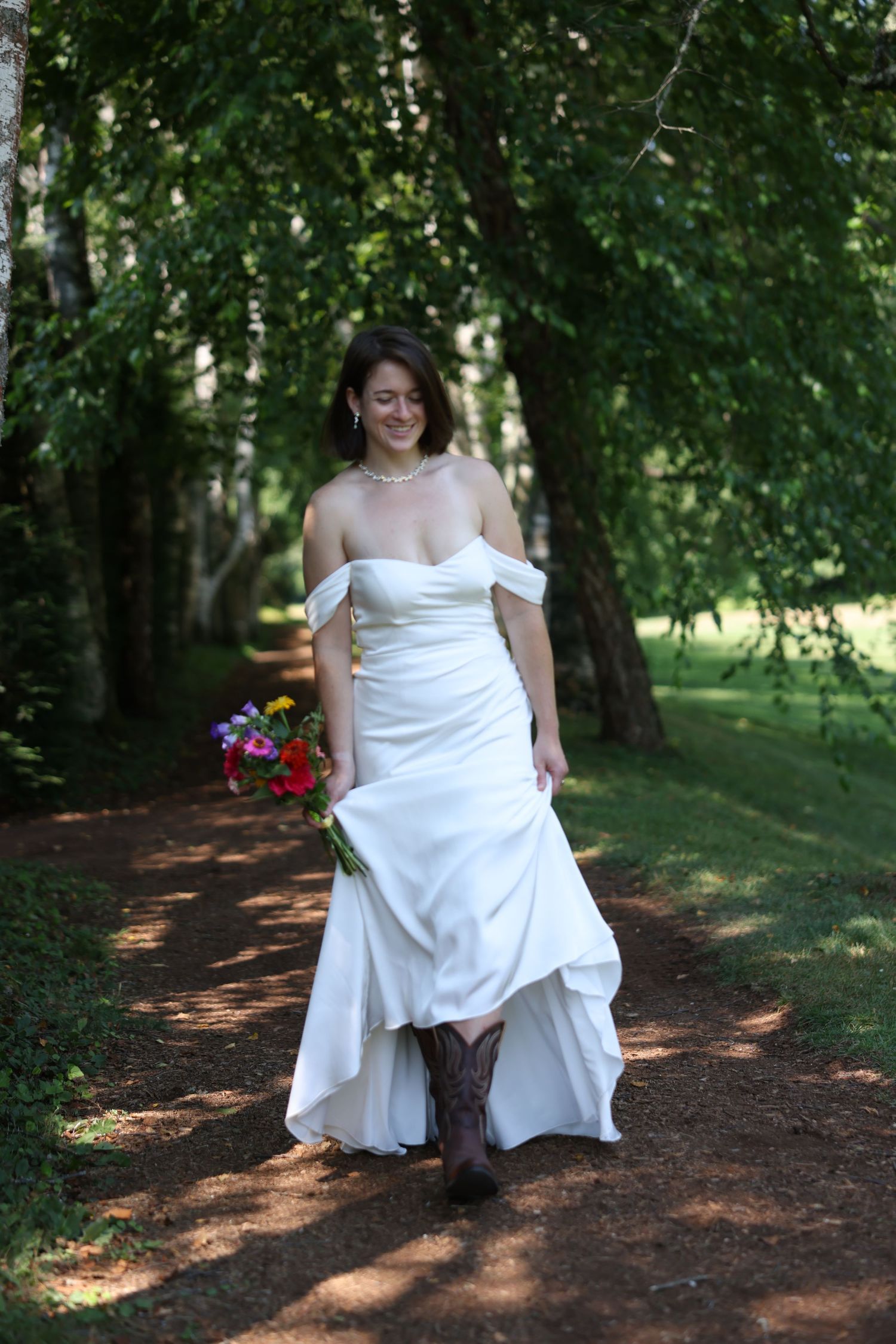Bride in white strapless wedding dress and brown cowboy boots walking along a tree-lined dirt path with bouquet.