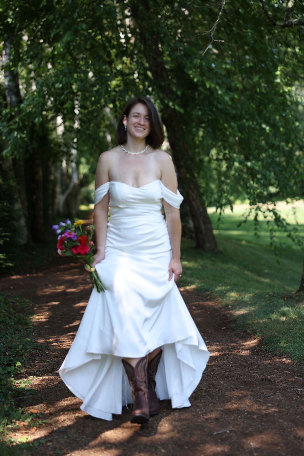 Smiling bride in white off-shoulder gown and cowboy boots holding colorful bouquet on a shaded garden path.