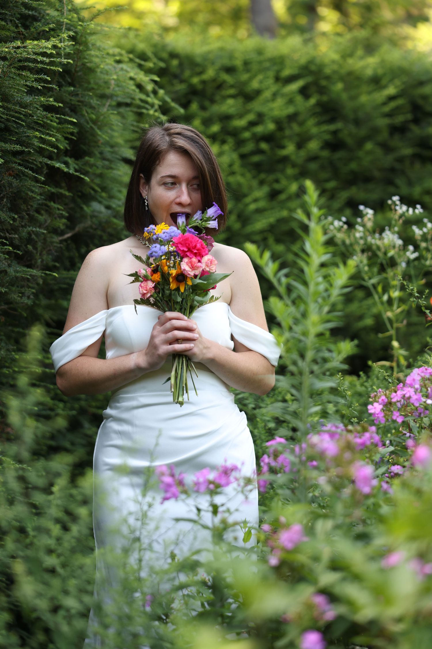 Bride in white off-shoulder dress smelling a vibrant wildflower bouquet surrounded by lush garden greenery.
