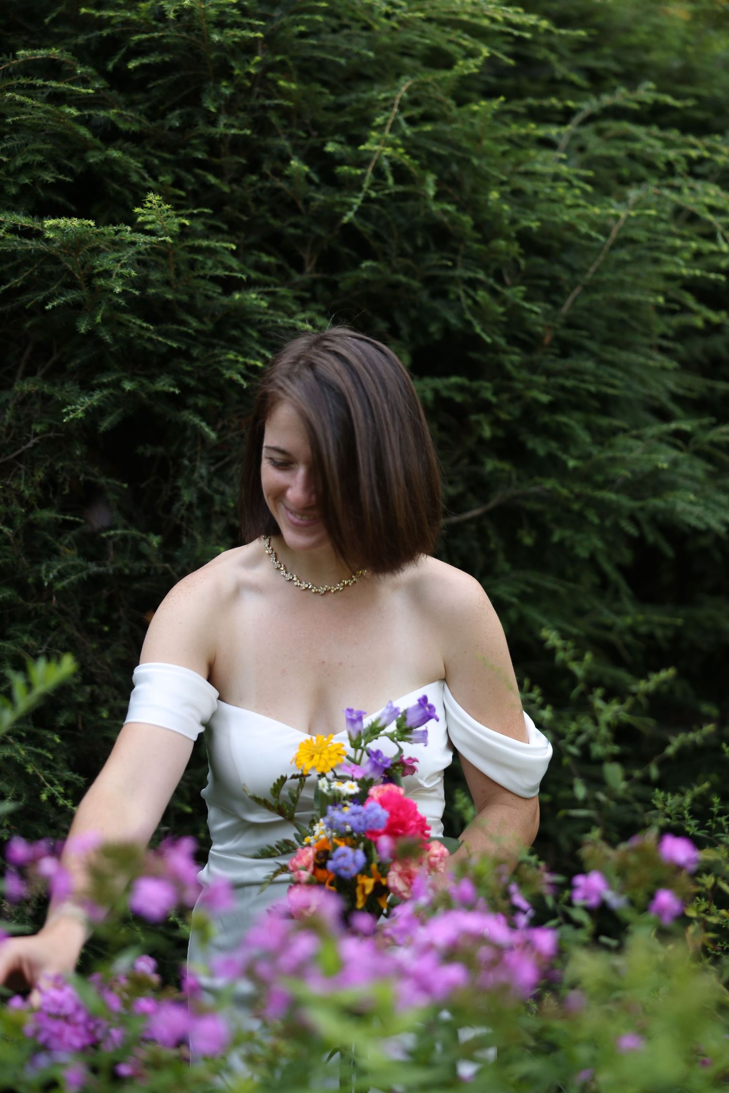 Bride in white strapless gown with gold necklace holding colorful bouquet among purple flowers and green trees.