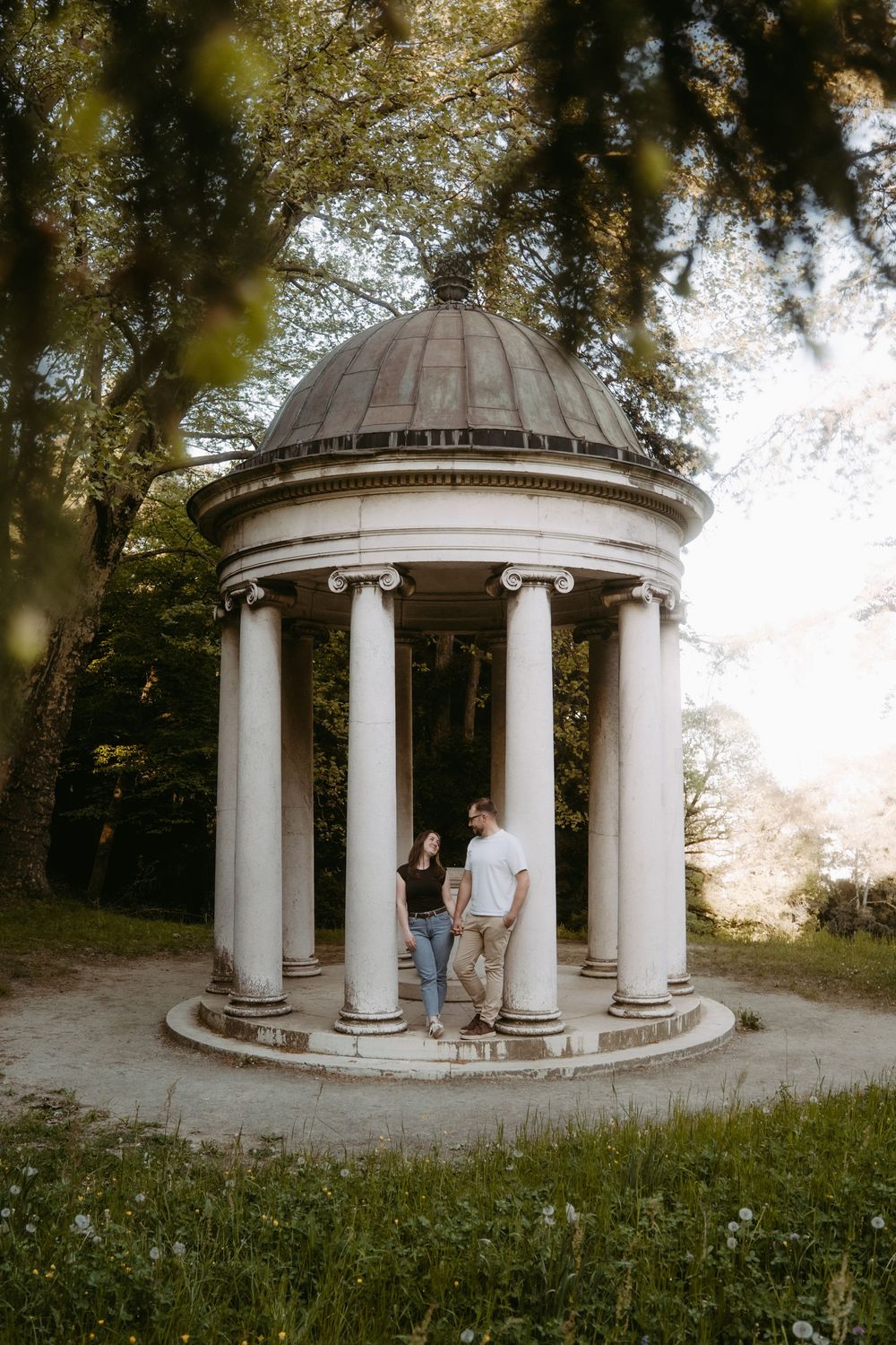 Couple standing together under a classical white domed rotunda with columns in a lush green park setting.