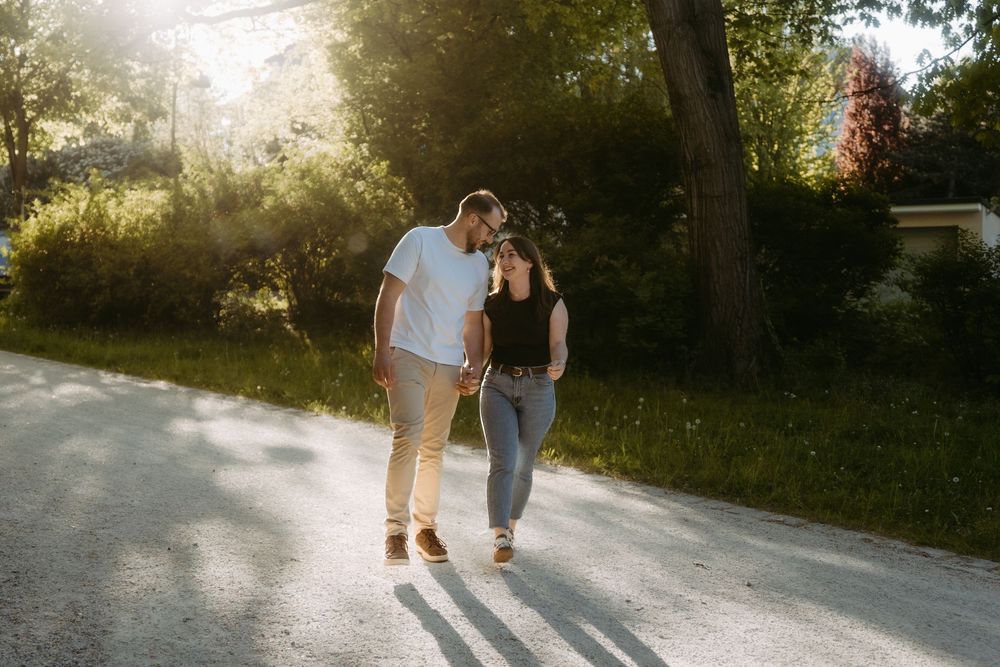 Couple walking hand in hand on a sunlit path surrounded by lush green trees during golden hour.