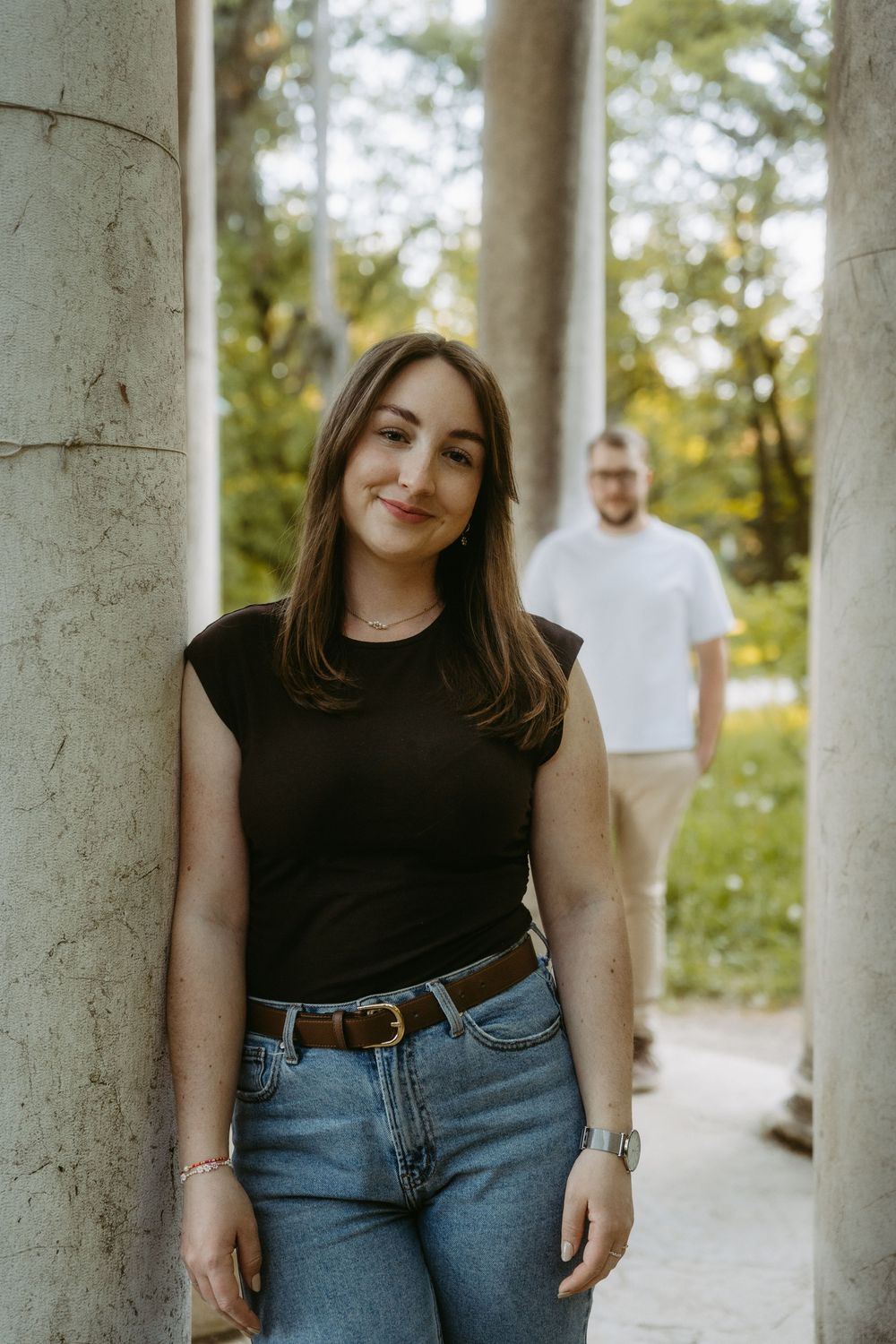 Young woman in black t-shirt and jeans leaning against stone column in a park, man standing in background.