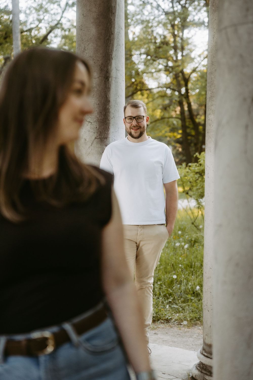 A smiling man in white t-shirt and glasses stands between stone columns in a park, while a woman in black top stands in foreground.