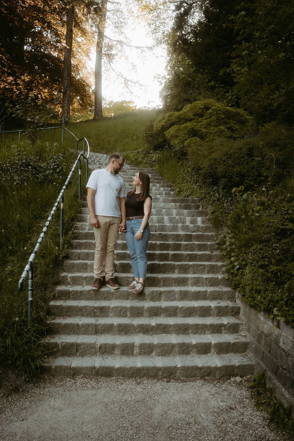 Couple holding hands on stone steps surrounded by lush green trees at golden hour.