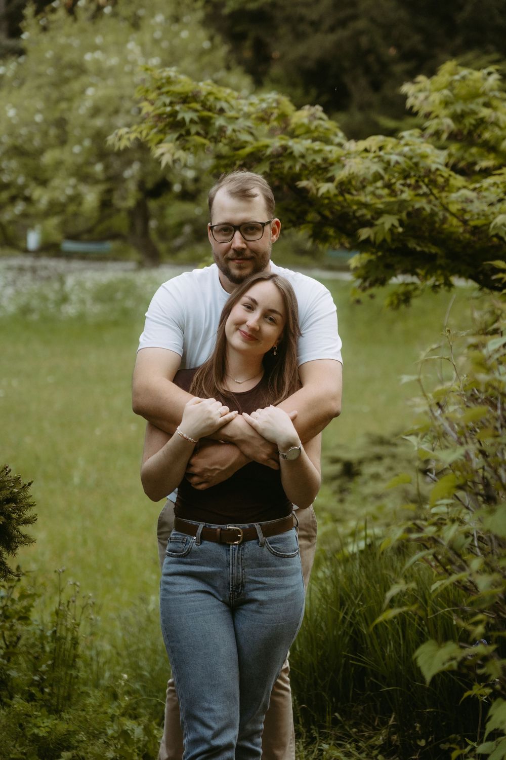 A couple embraces in a lush green garden, man in white tee hugging woman in brown top and jeans.