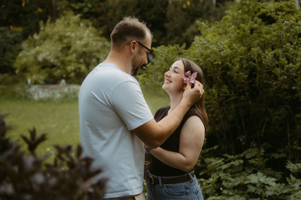 A couple shares a tender moment outdoors, man gently tucking hair behind womans ear amid lush green garden backdrop.