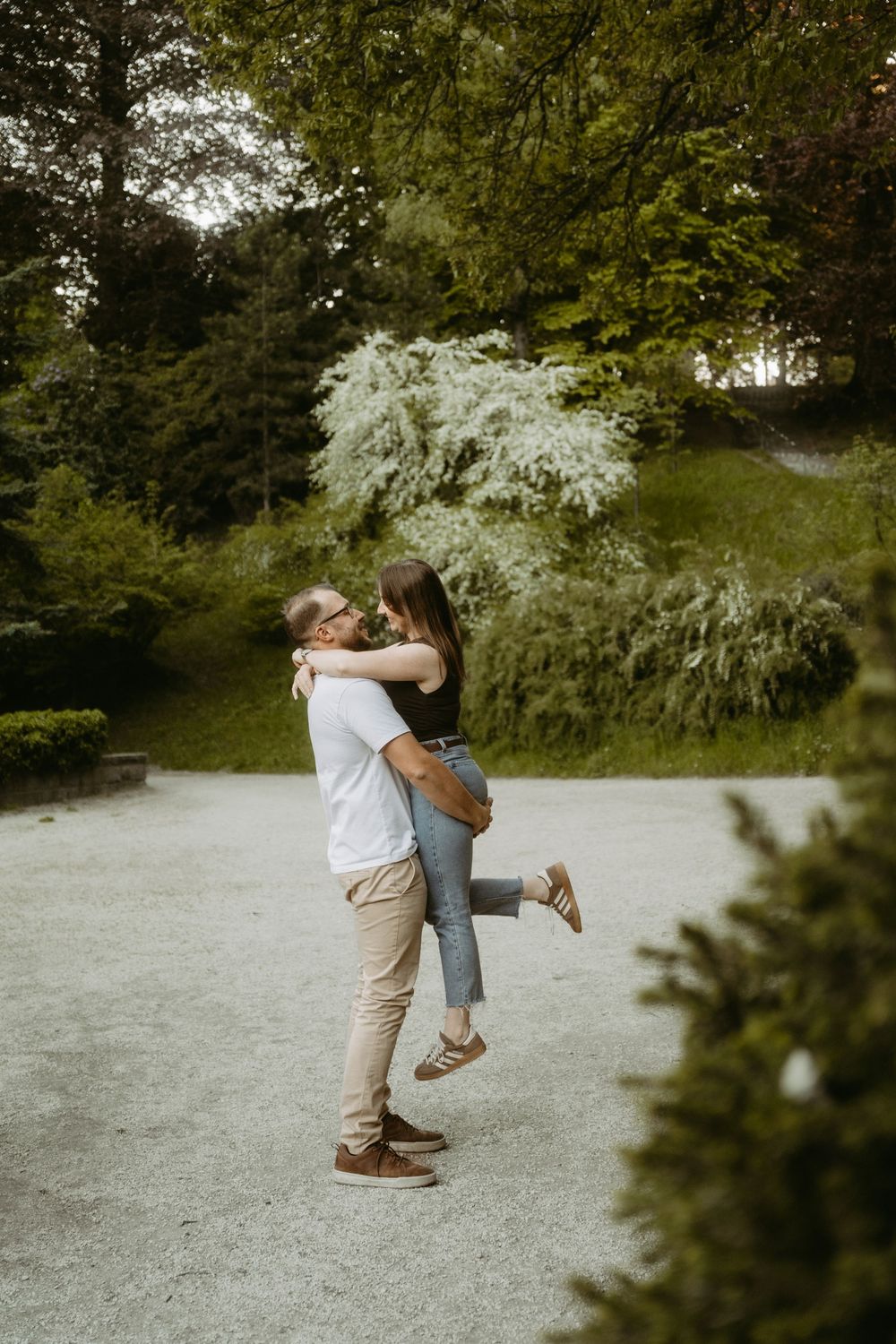 Man lifting woman in a joyful embrace on a garden path surrounded by lush green trees.