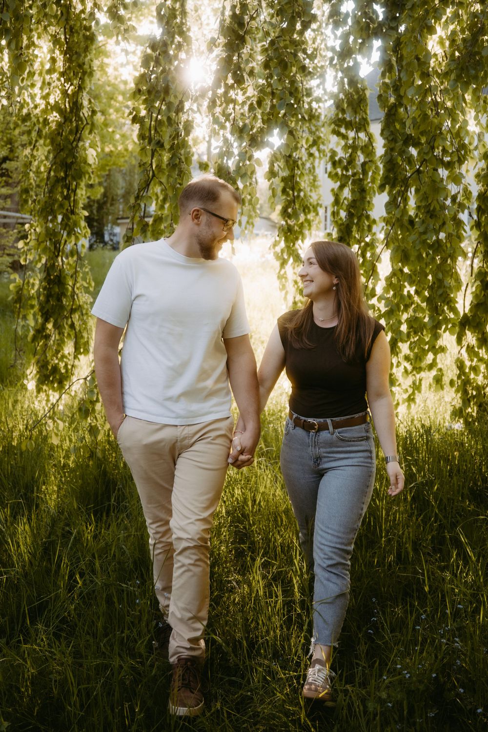 Couple holding hands walking through sunlit park with lush green trees, smiling at each other on a warm summer day.