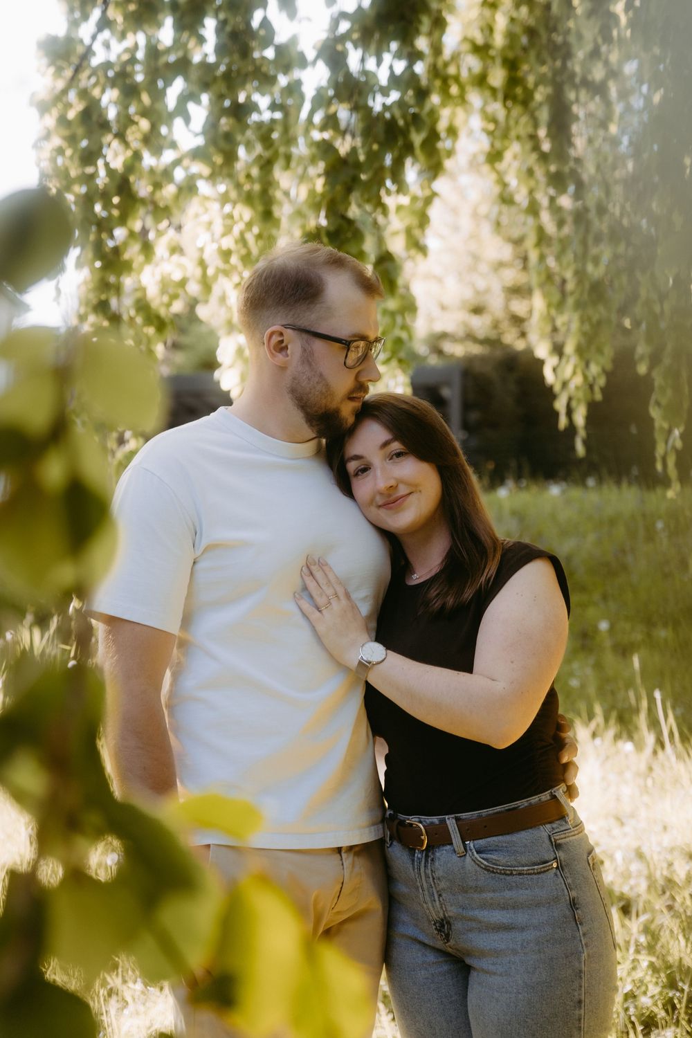 A couple embraces tenderly outdoors under a willow tree in warm sunlight, man in white tee, woman in black top and jeans.