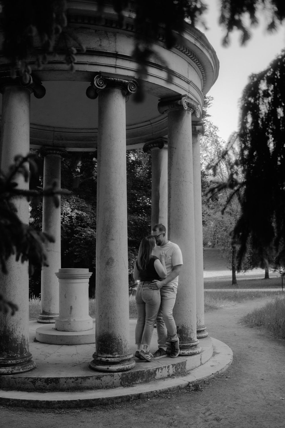 Black and white photo of a couple embracing between classical columns of a rotunda in a park setting.