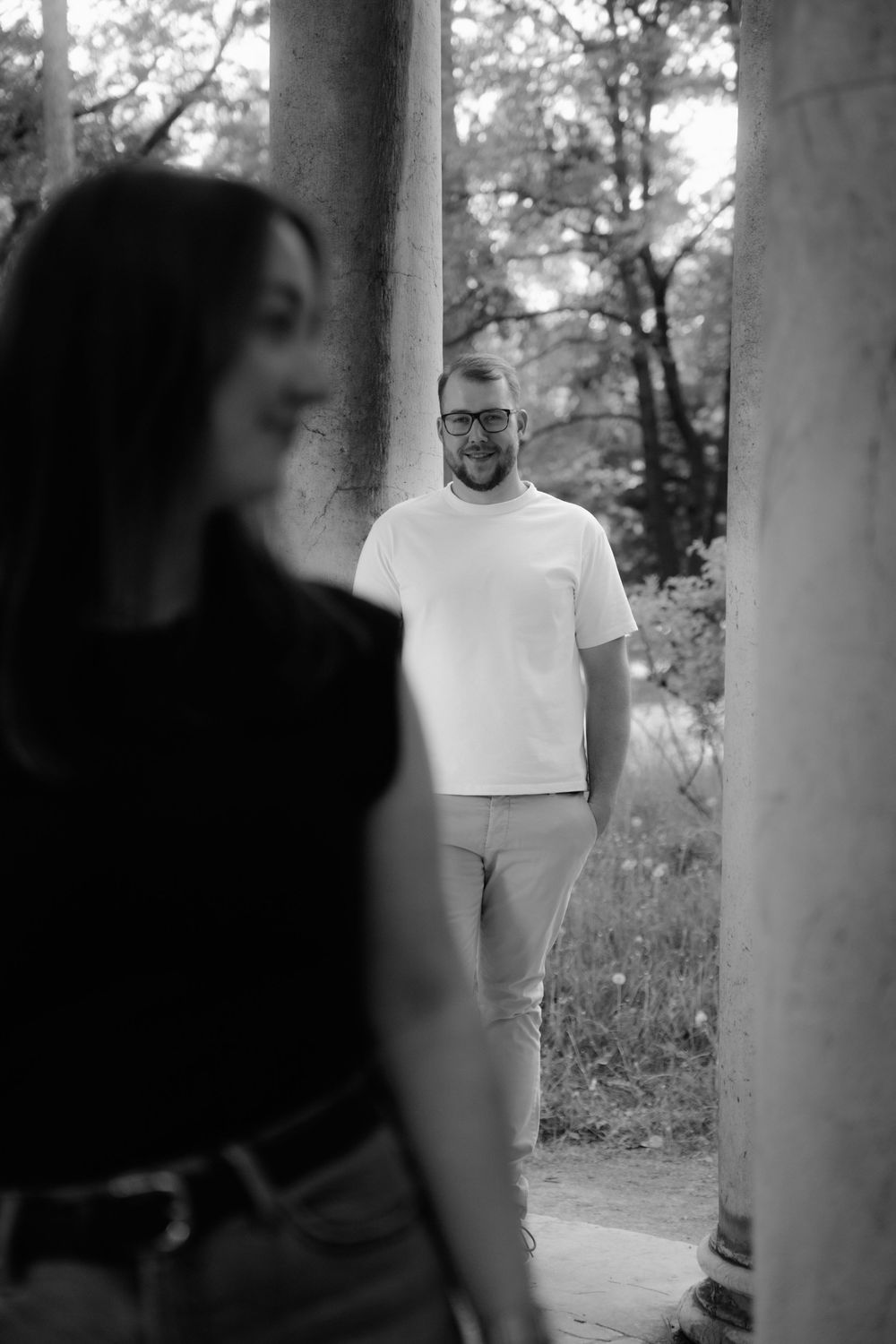 Black and white photo of a smiling man in white t-shirt standing between columns, with a woman in foreground.