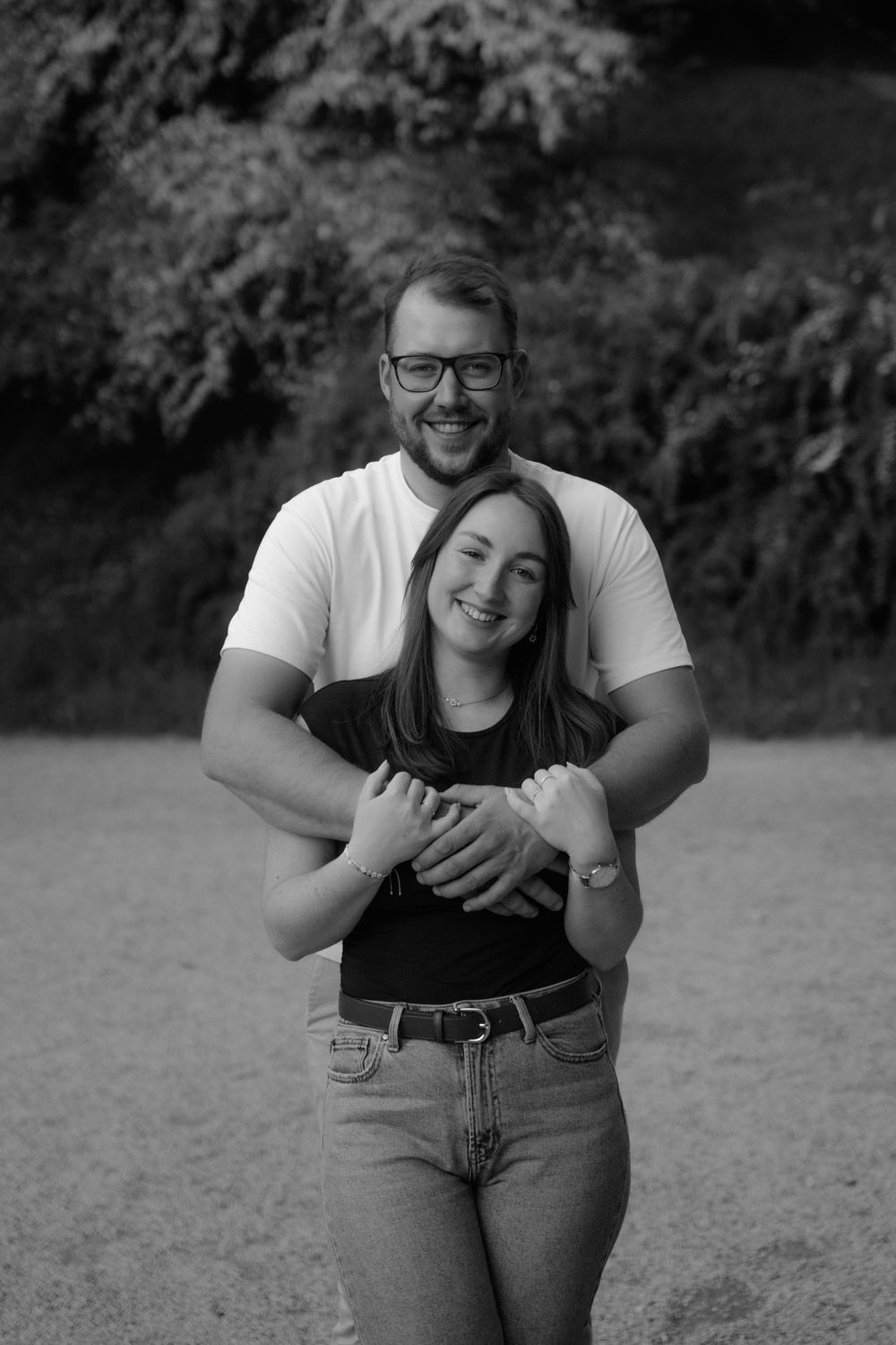 Black and white photo of a smiling couple embracing outdoors, man in glasses hugging woman from behind near trees.