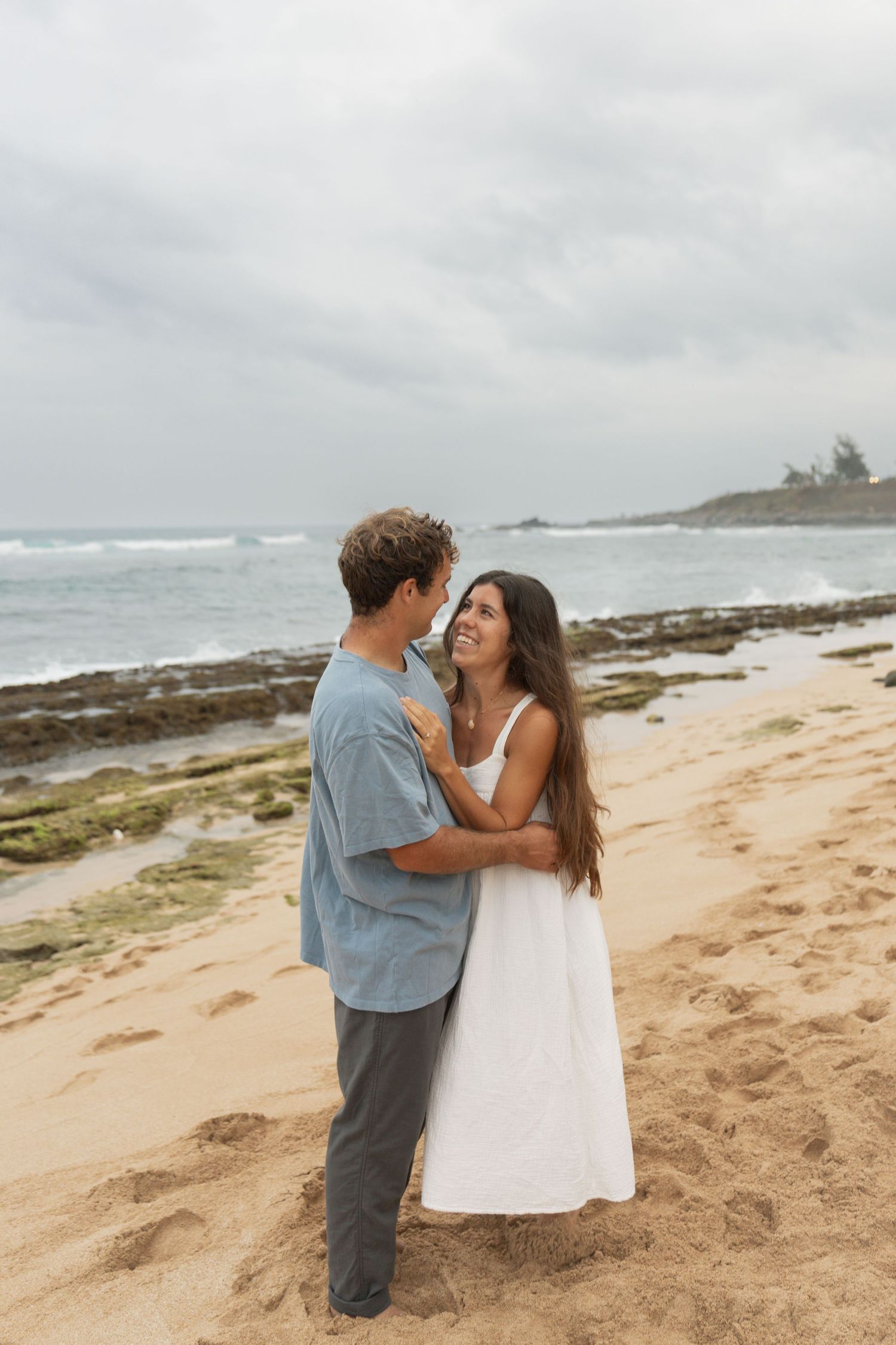 Couple holding hands on a coastal cliff with ocean waves and rocky shoreline in the background on an overcast day.