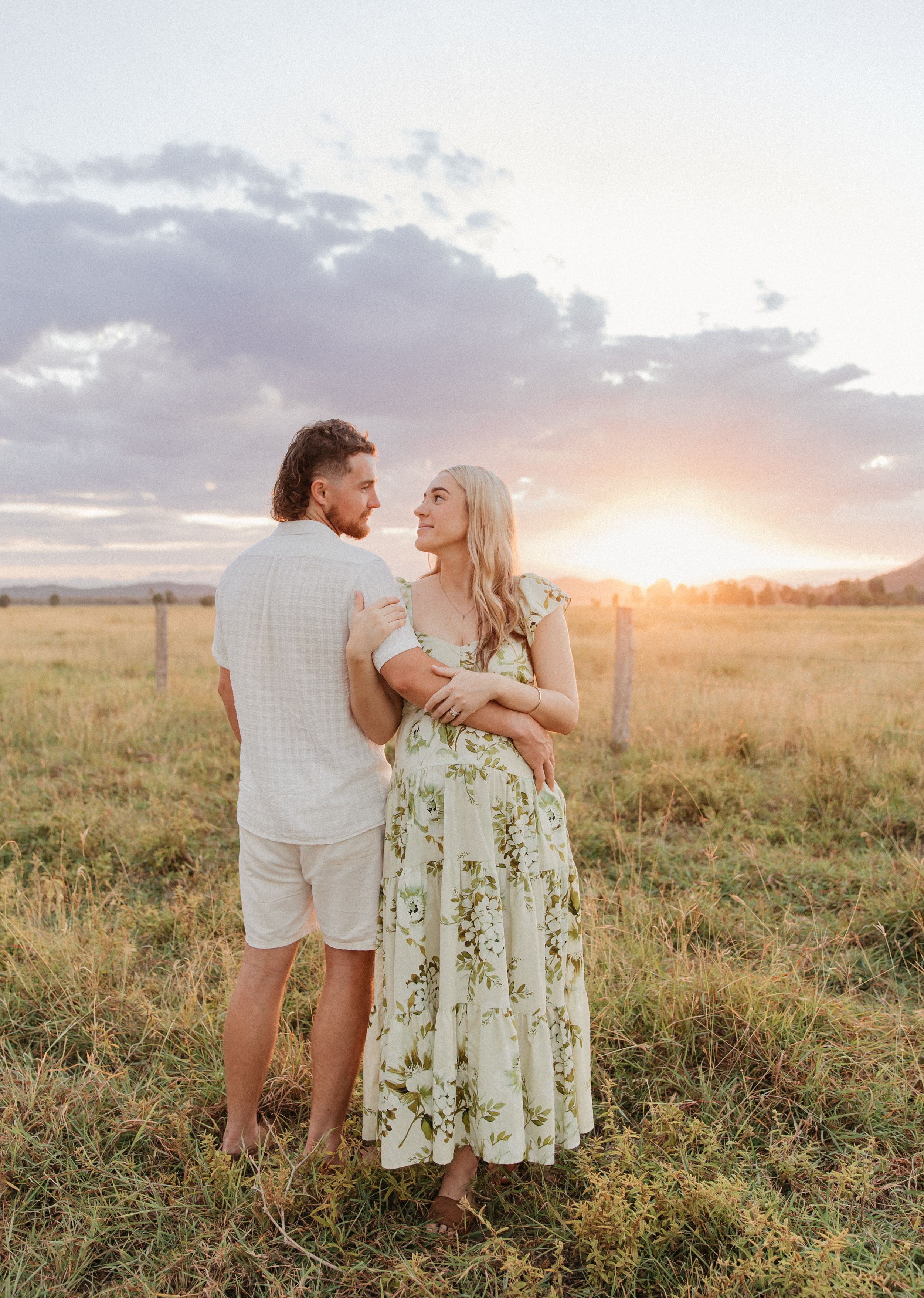 Couple embracing in a golden field at sunset, woman in floral dress and man in white shirt and shorts.