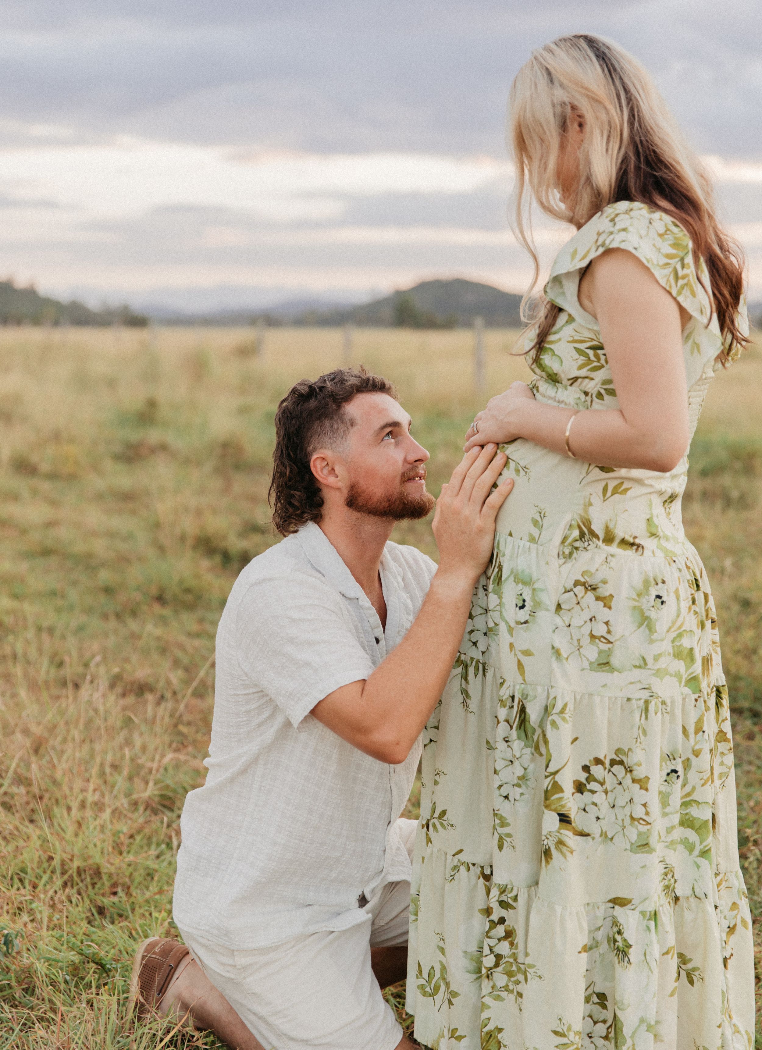A man kneels in a field touching a pregnant woman's belly in a romantic outdoor maternity photo shoot.