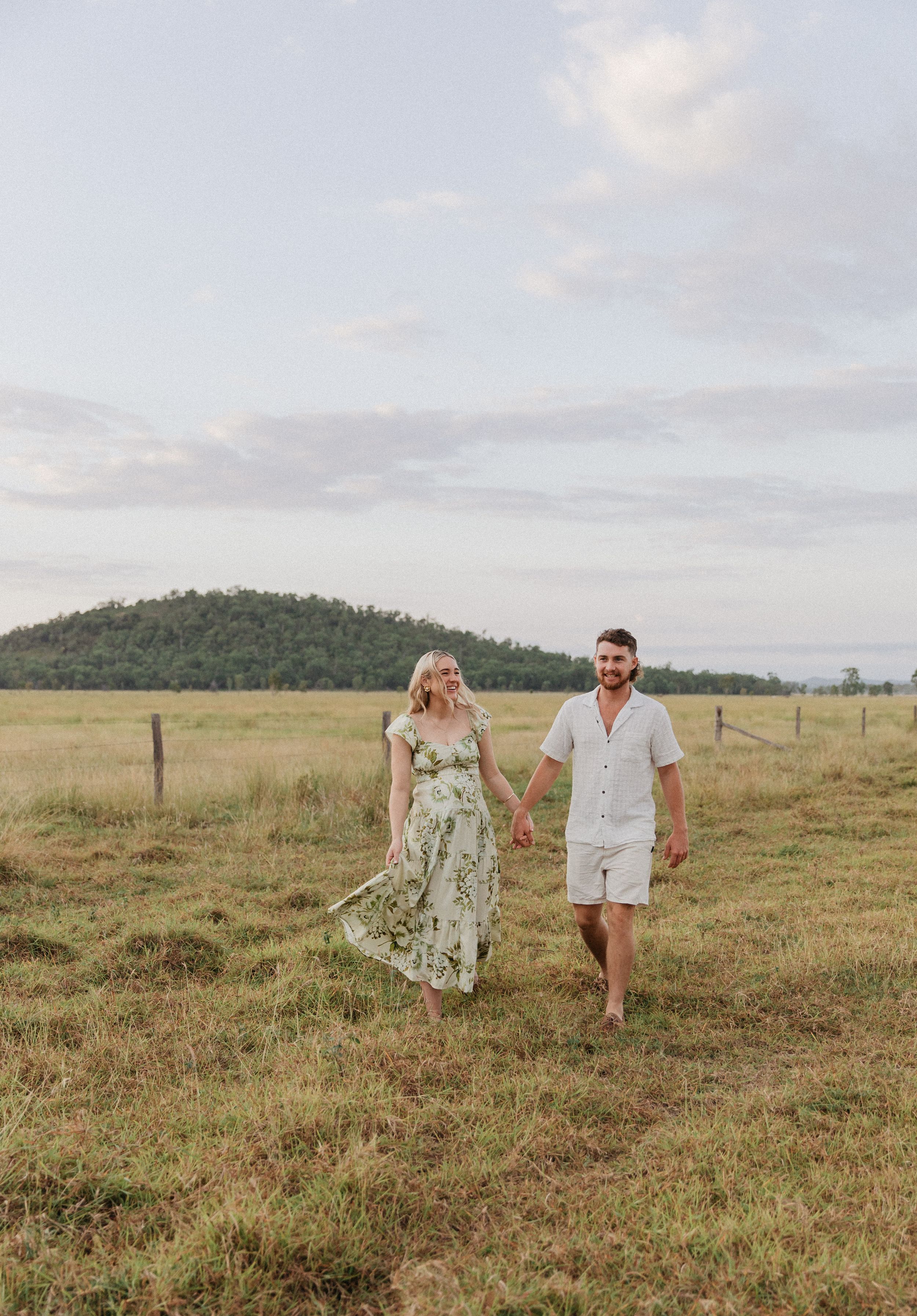 Couple holding hands walking through a green field with a hill and cloudy sky in the background.