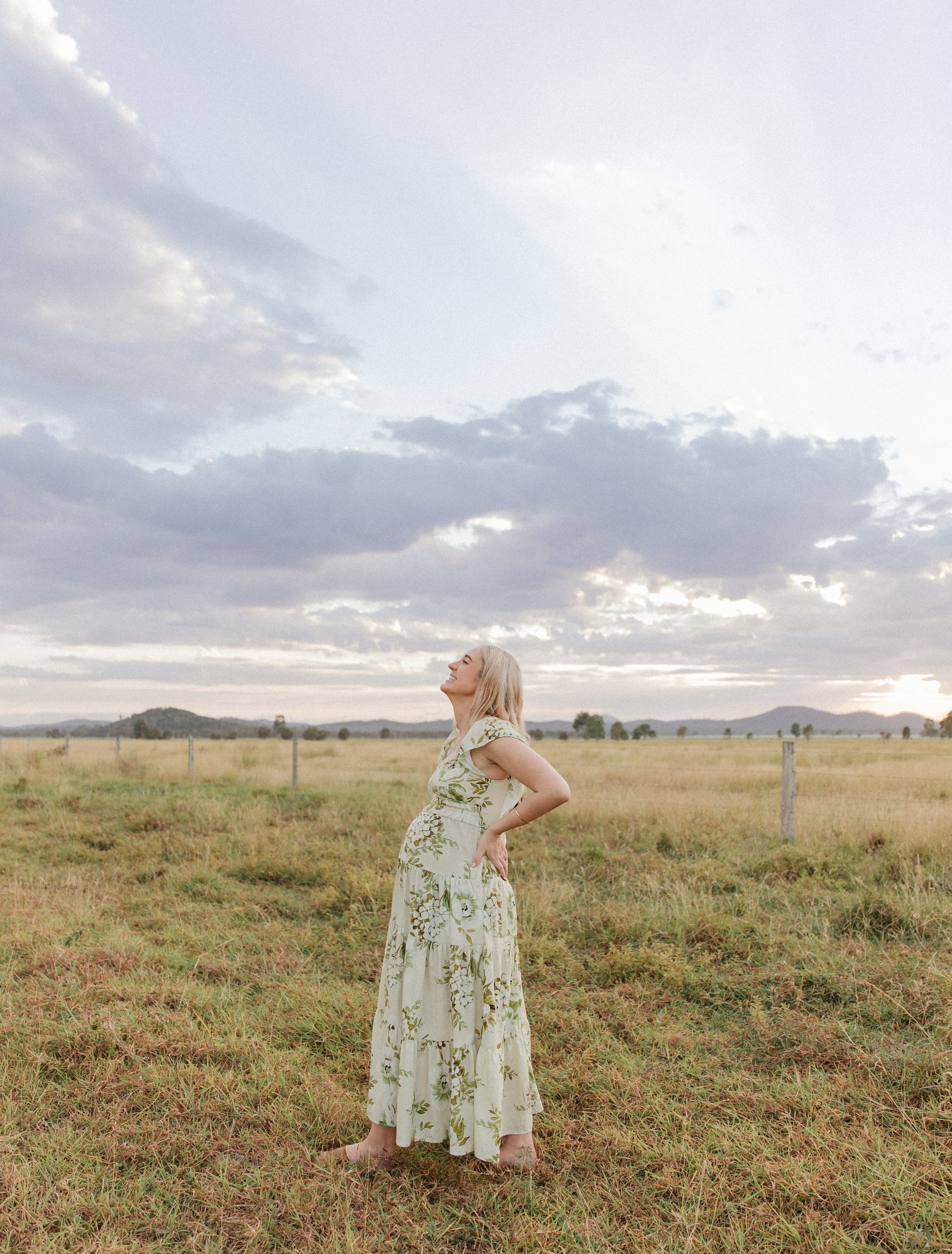 Blonde woman in floral green dress standing in open field looking up at dramatic cloudy sky at sunset.