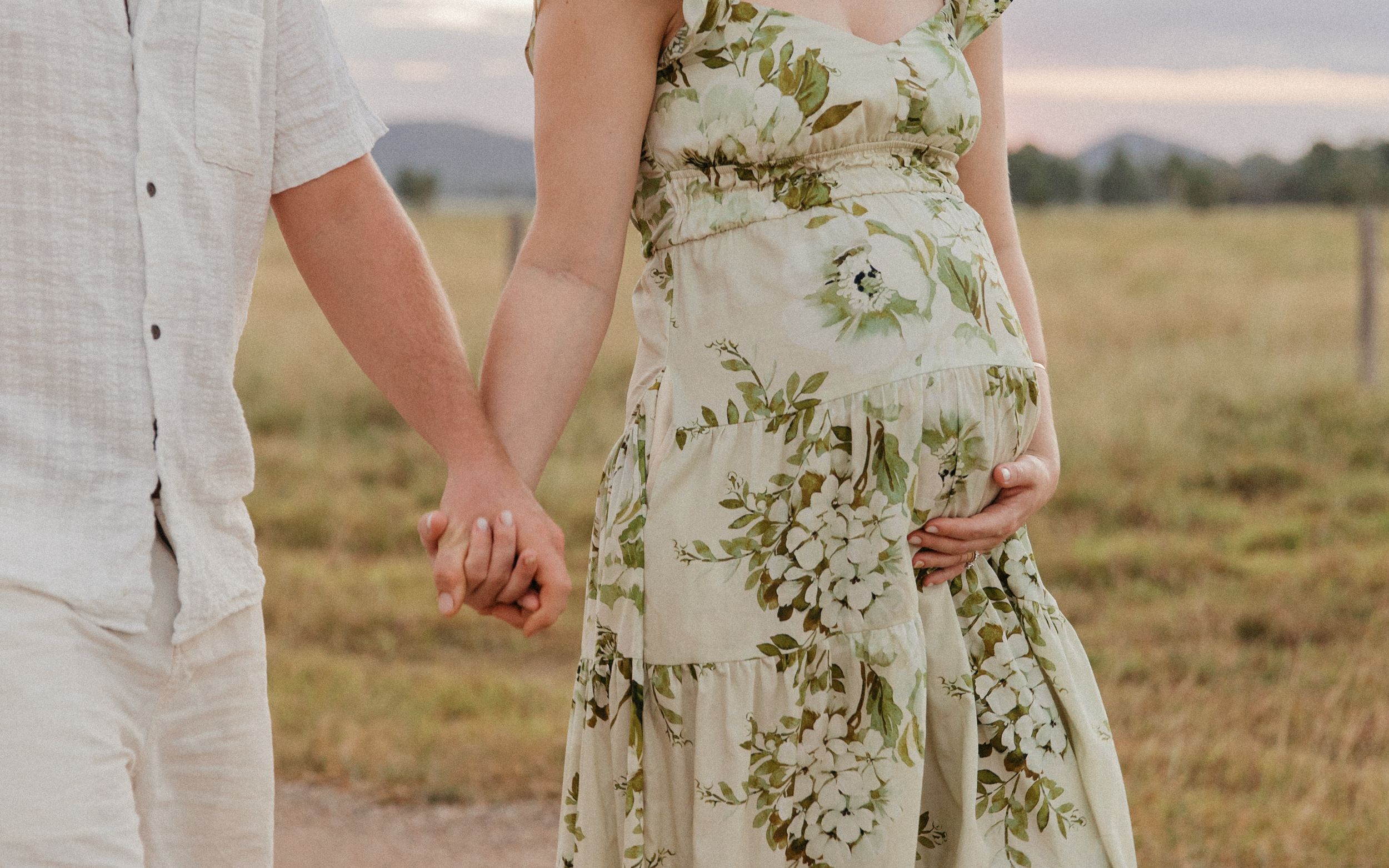 Pregnant couple holding hands in a field, woman wearing a floral green maternity dress, cradling her baby bump.