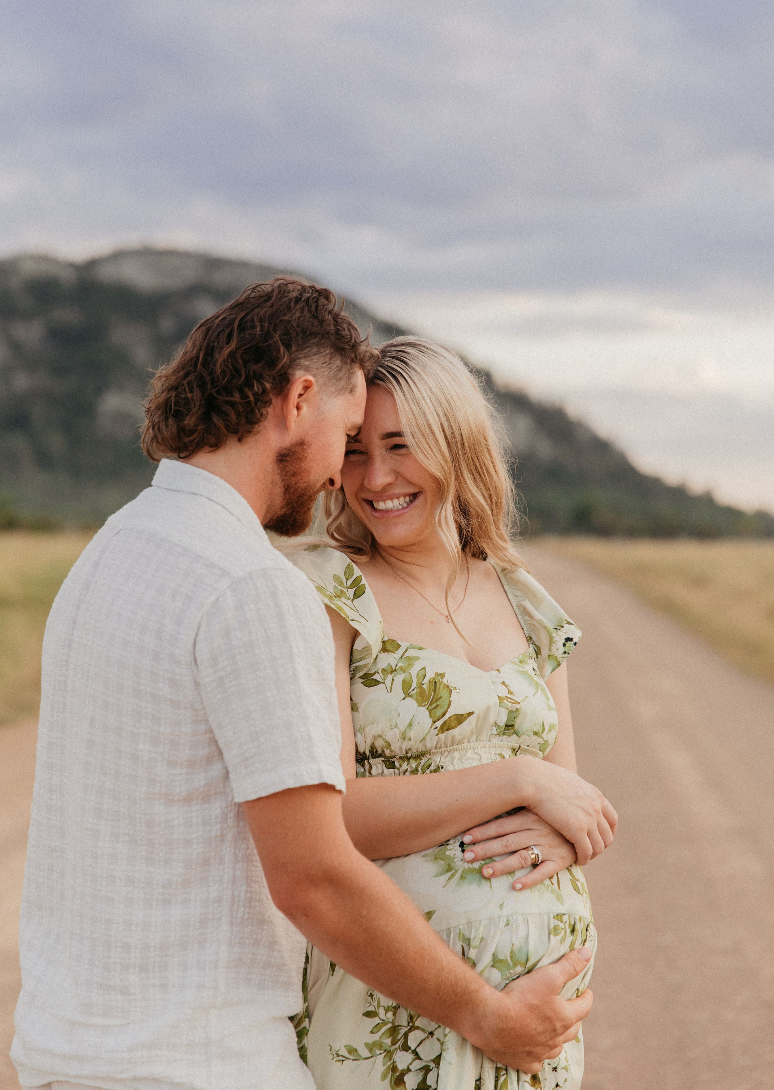 Couple embracing on a dirt road with mountains, man nuzzling pregnant woman in floral dress, outdoor maternity photo.