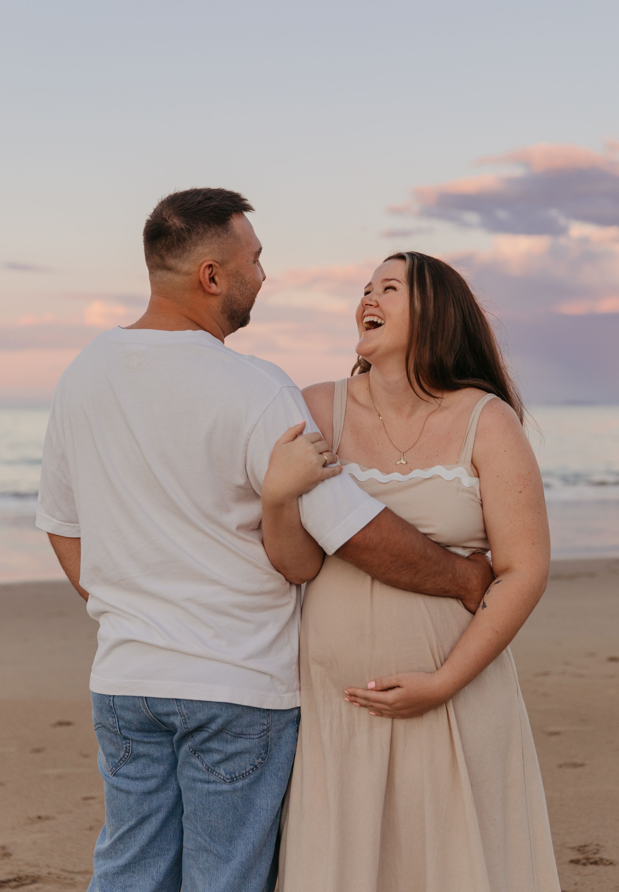 Pregnant couple laughing together on a beach at sunset, woman in beige dress cradling her baby bump.