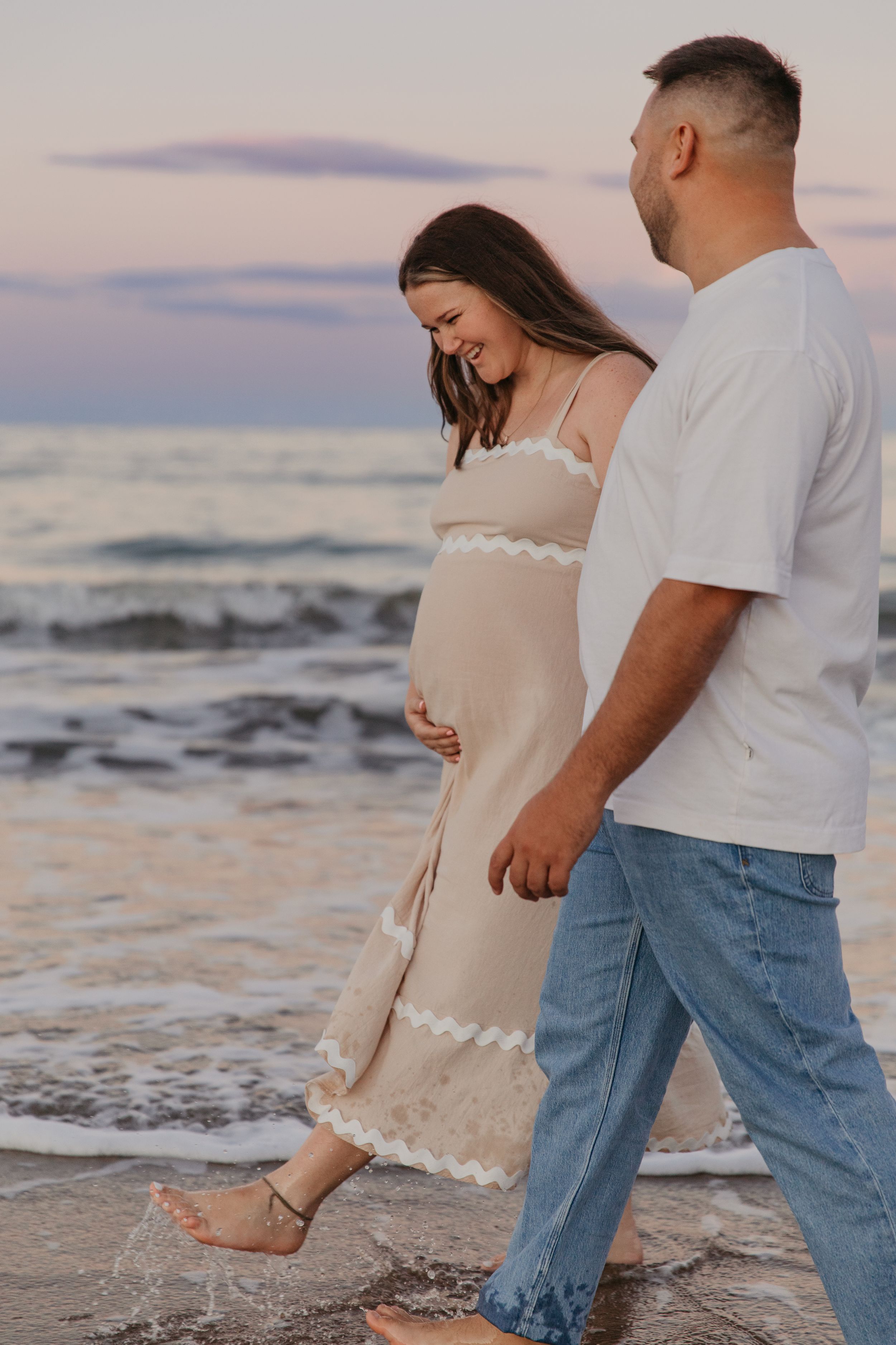 Pregnant couple walking barefoot on beach at sunset, woman in beige dress holding baby bump, man in white tee and jeans.