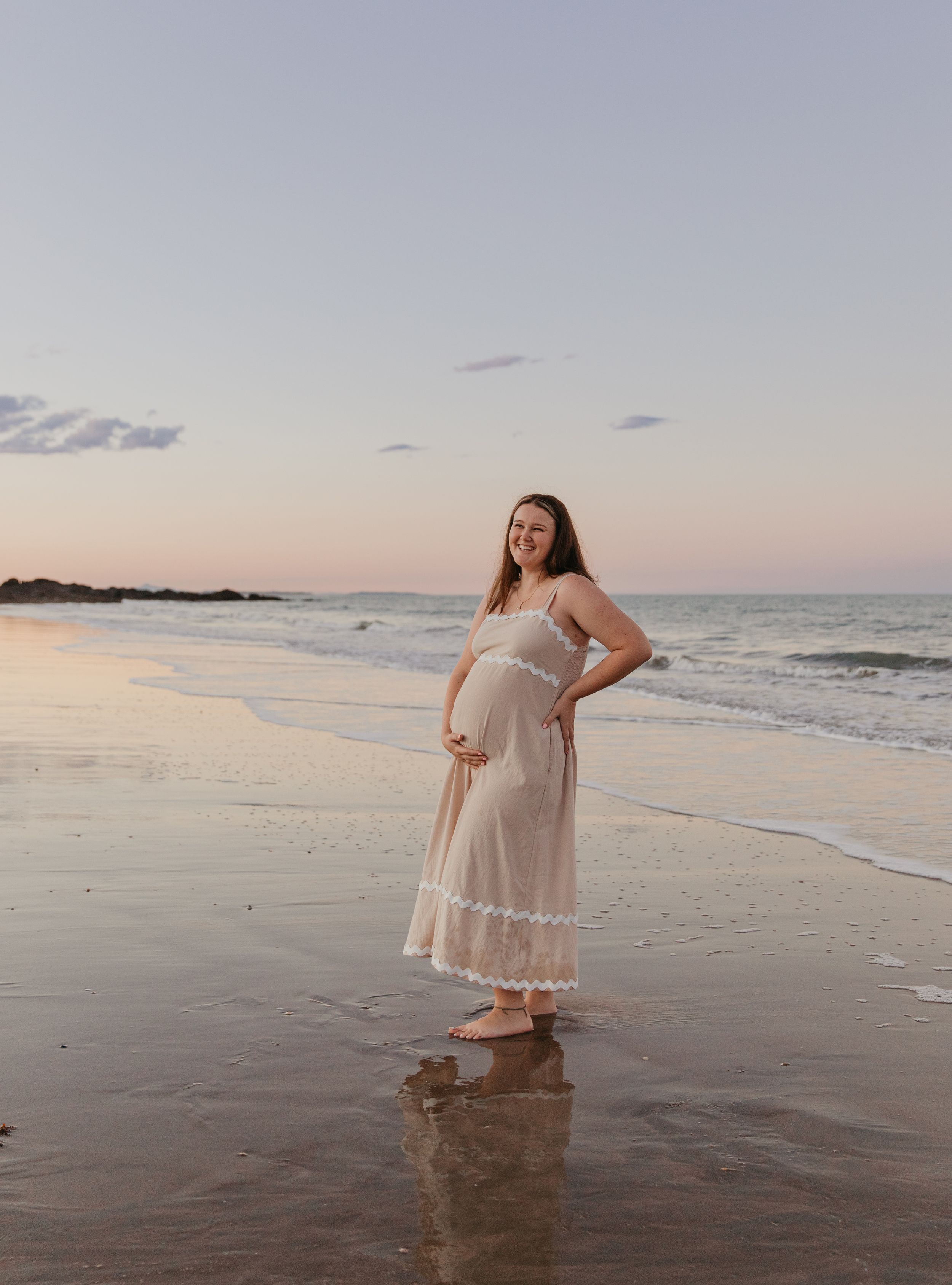 Pregnant woman in white lace dress smiling on a beach at sunset, cradling her baby bump near ocean waves.