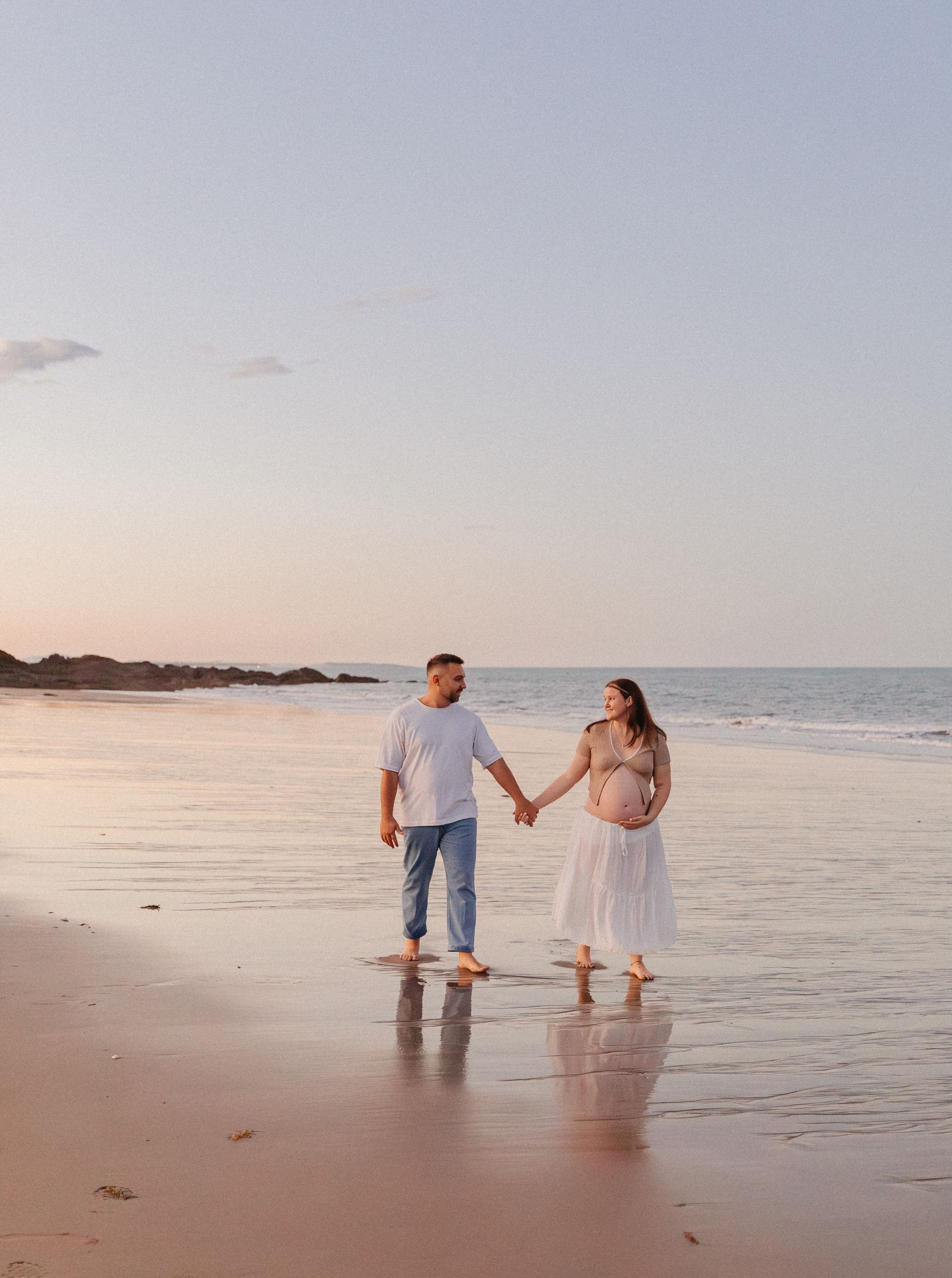 Pregnant couple holding hands on a sandy beach at sunset, woman in white dress, man in jeans and white shirt.