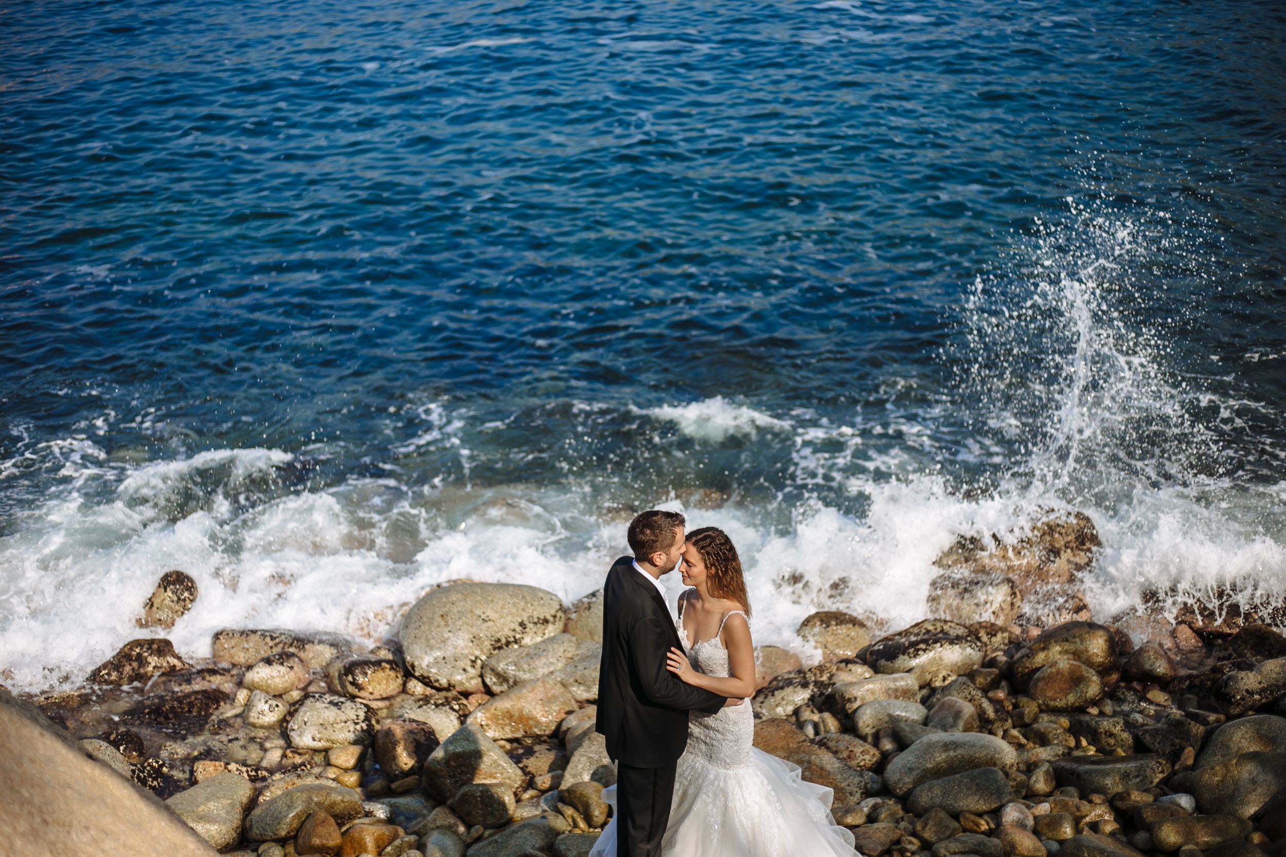 Couple embraces on rocky coastline as waves crash against shore during sunset photo session.
