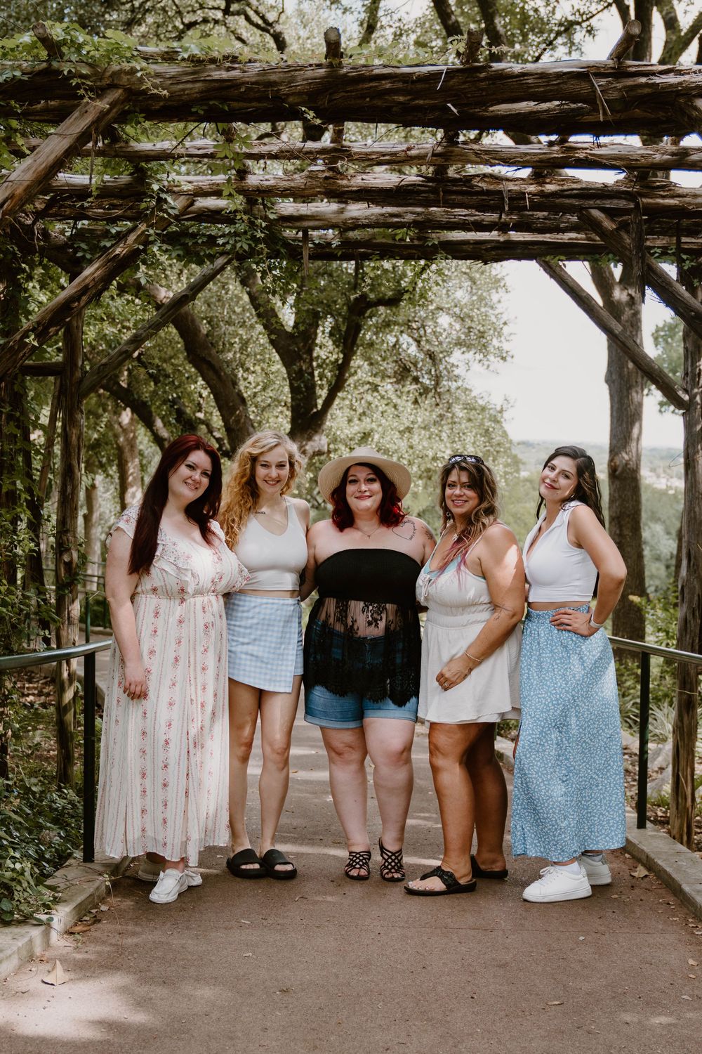 Five women posing together under a rustic wooden pergola surrounded by lush green trees on a sunny day.