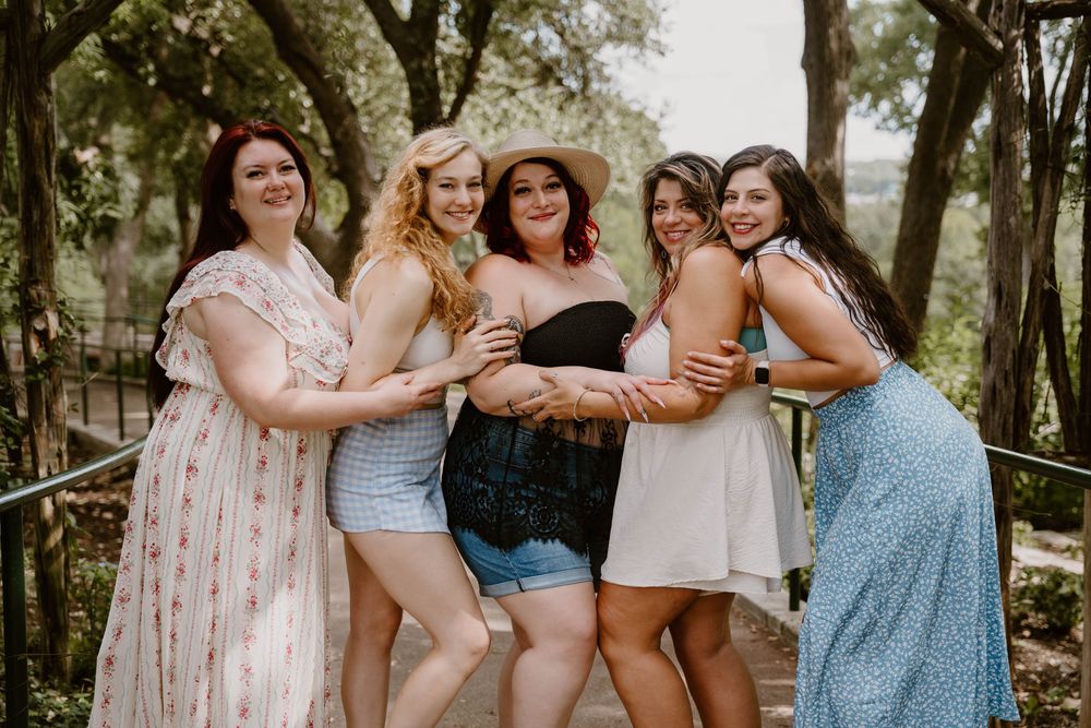 Five women posing together outdoors, smiling and embracing, wearing summer dresses and casual outfits in a lush green setting.