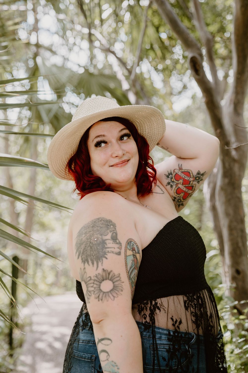 Tattooed woman with red hair and straw hat posing outdoors among tropical trees in black top and denim.