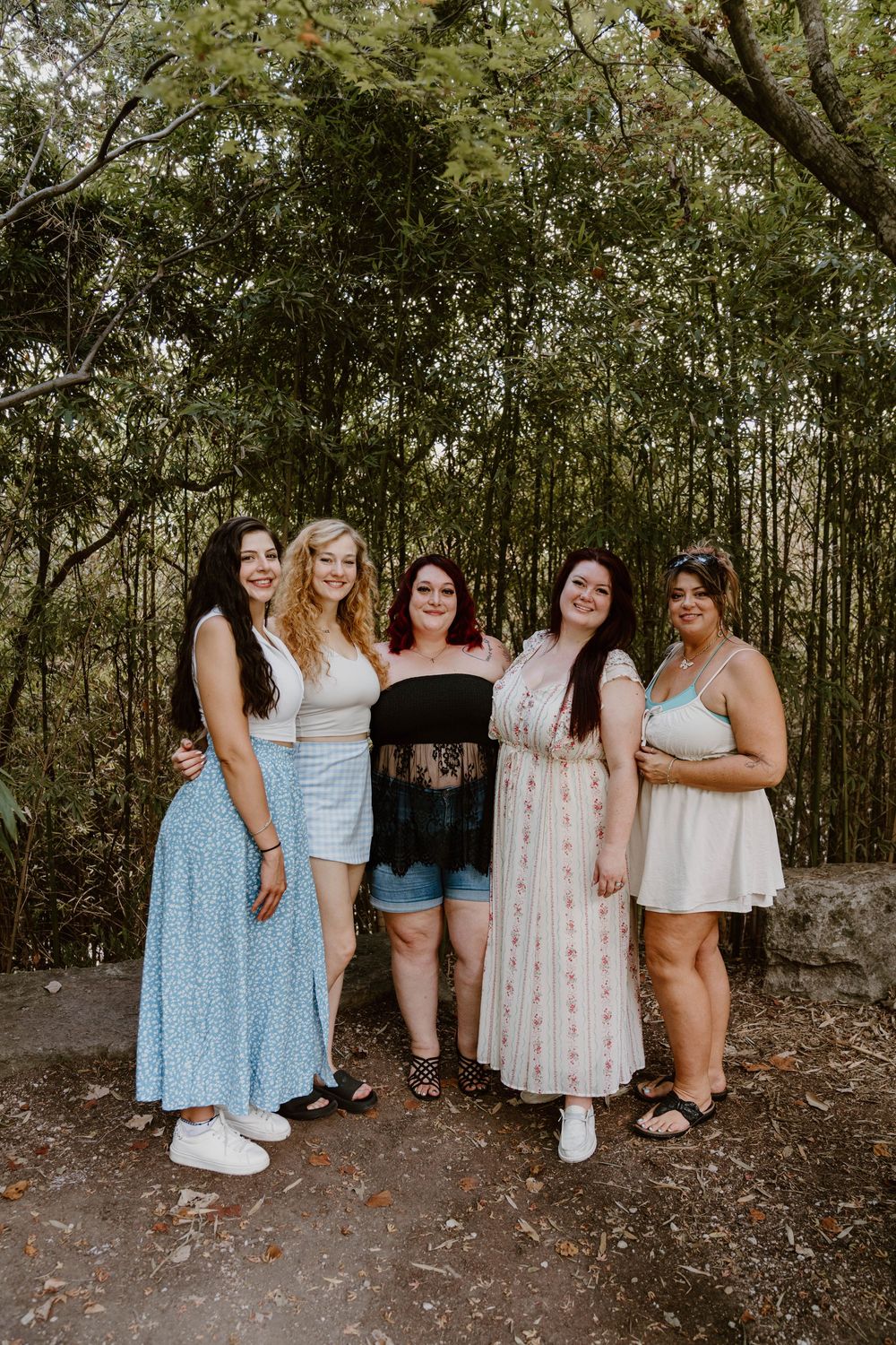 Five young women smiling and posing together outdoors in front of lush green bamboo trees on a sunny day.