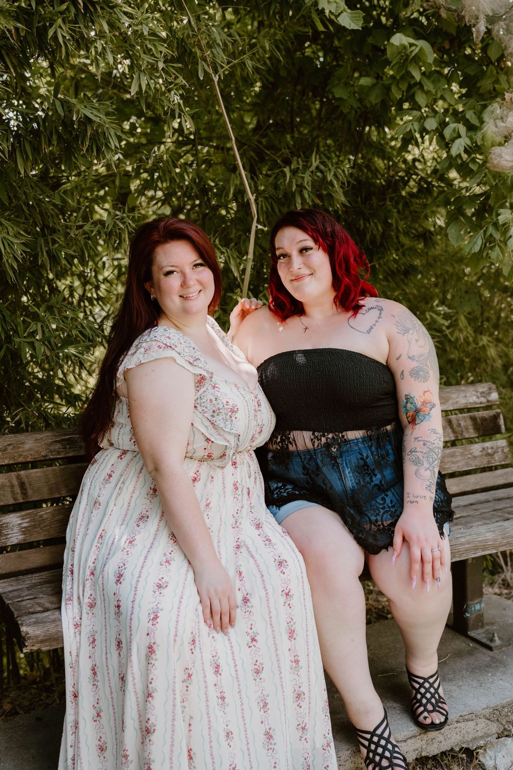 Two women with red hair sit together on a wooden bench outdoors, one in a floral dress, the other in a black top and denim shorts.