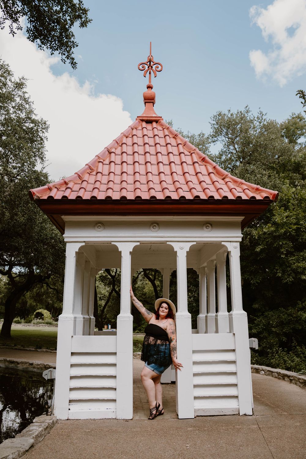 Woman posing in a white gazebo with red tile roof surrounded by lush green trees in a park setting.