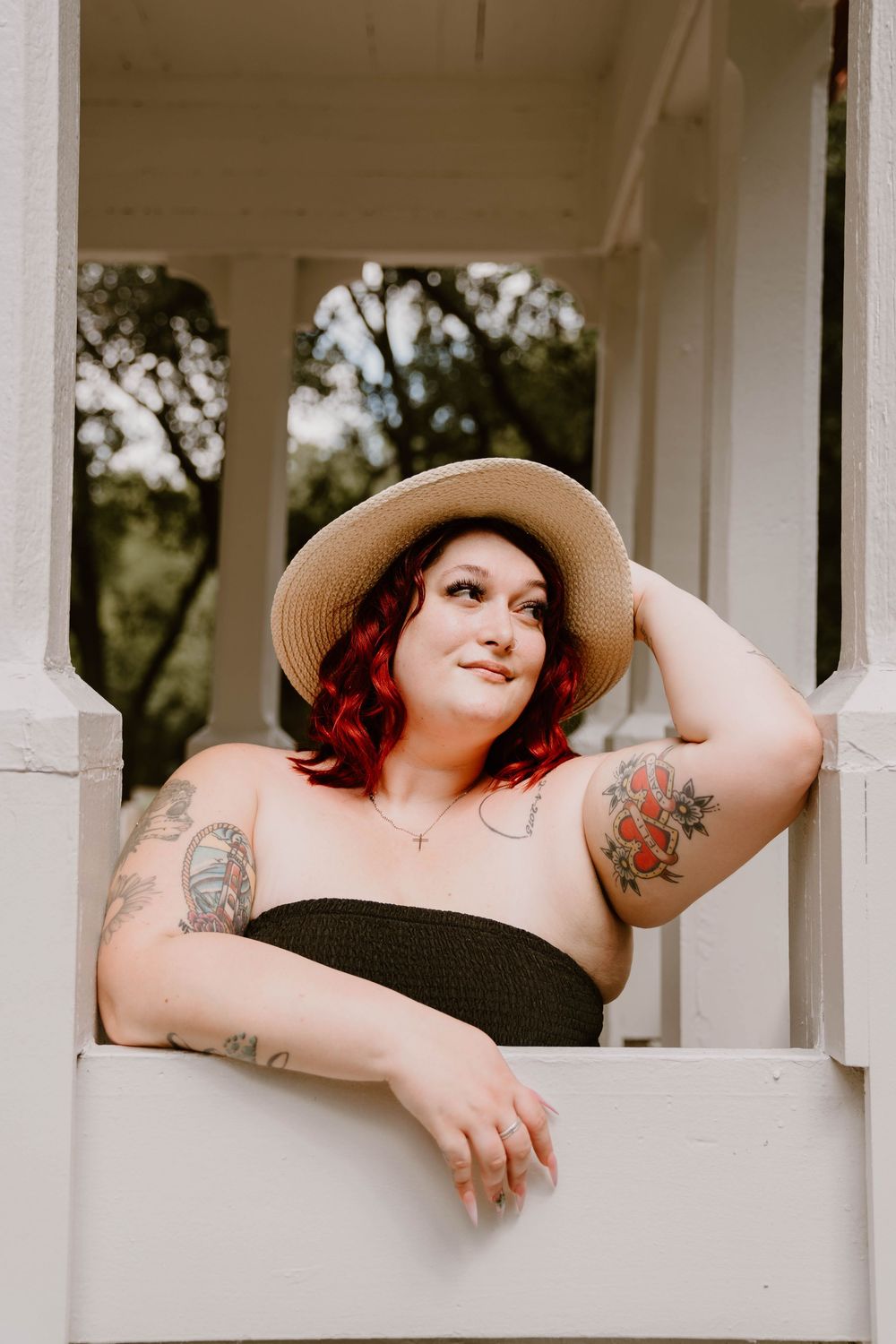 Red-haired woman with tattoos wearing straw hat and black tube top, leaning on white porch railing outdoors.