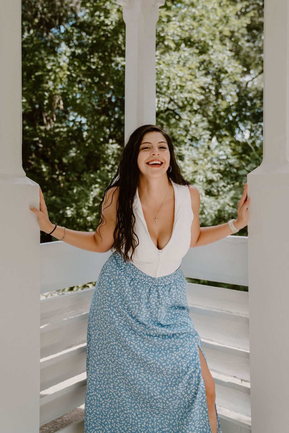 Smiling woman in white top and blue floral maxi skirt posing between white columns with green trees behind.