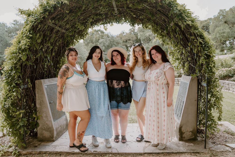 Five women posing together under a lush green floral arch in an outdoor garden setting.