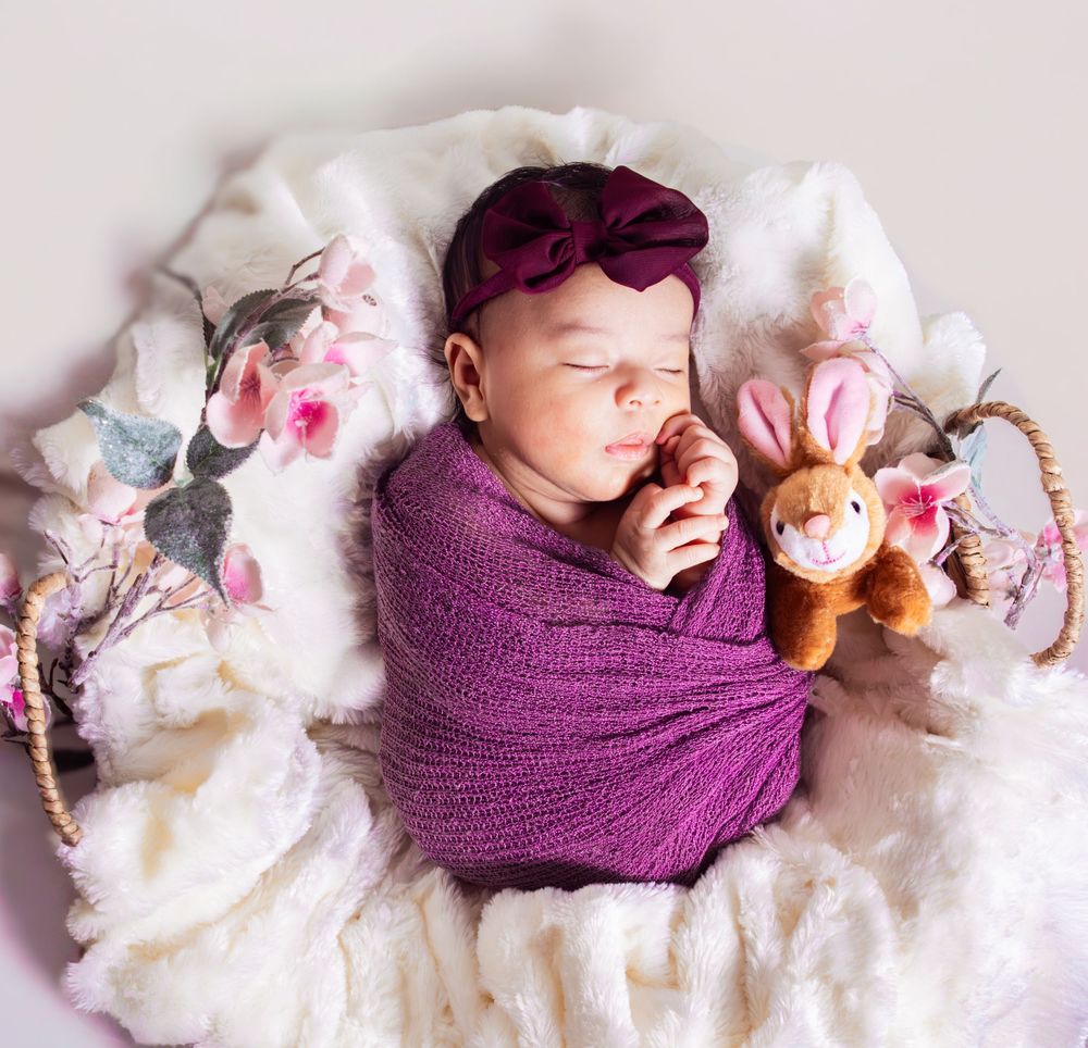 Sleeping newborn baby swaddled in purple wrap with bow headband in a basket with bunny stuffed animal.