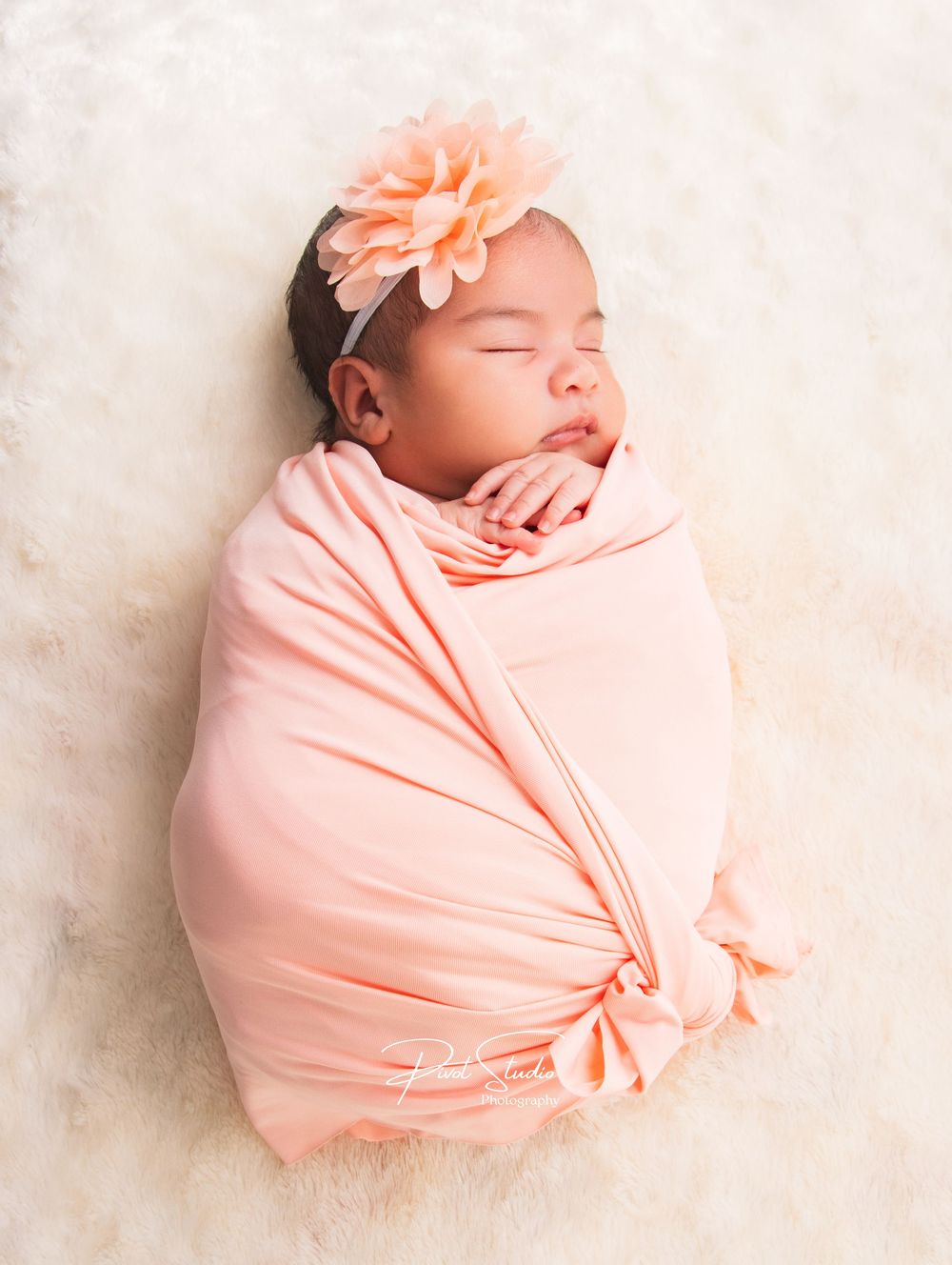Sleeping newborn baby swaddled in a soft peach wrap with a floral headband, lying on a fluffy white blanket.