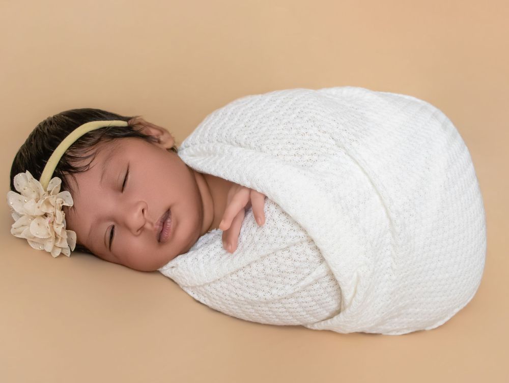 Sleeping newborn baby girl swaddled in white knit wrap wearing a delicate floral headband on beige background.
